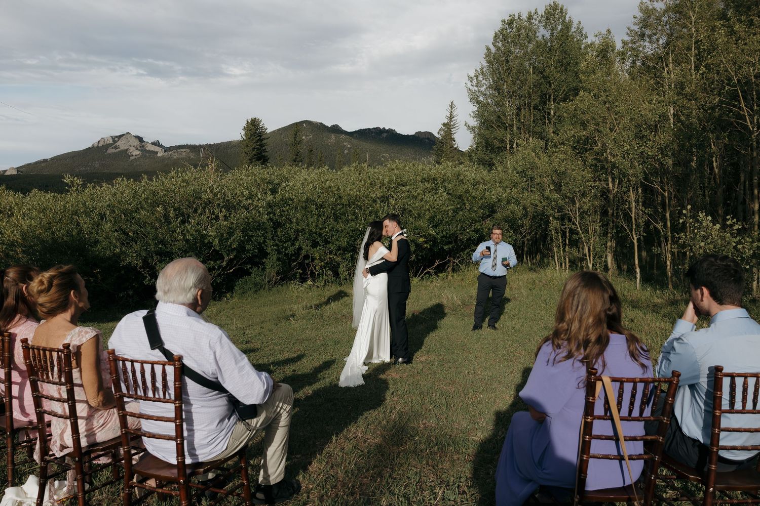 Couple sharing first kiss at outdoor mountain wedding ceremony with seated guests.