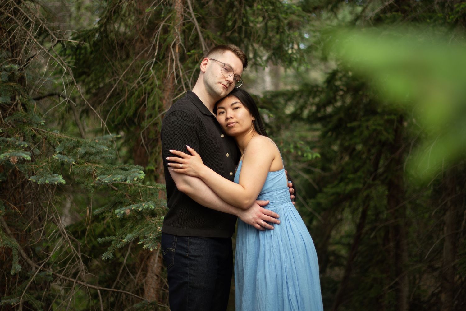 A couple shares a loving embrace among towering trees in natural lighting.