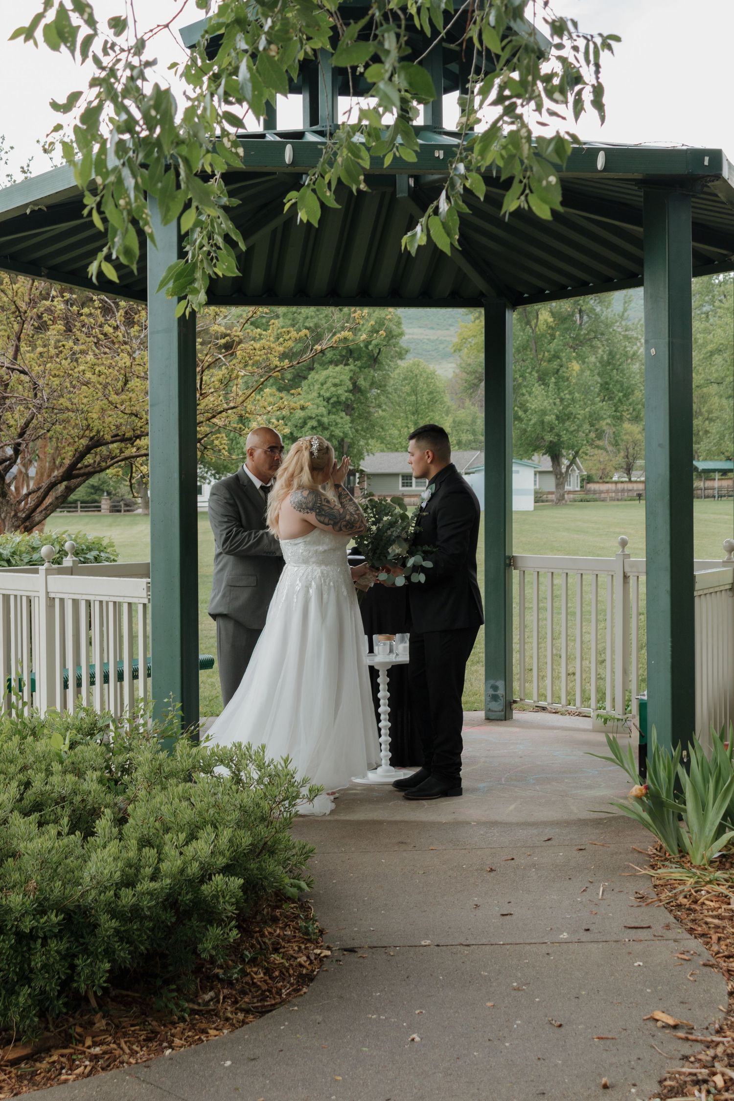 Wedding ceremony taking place under a covered outdoor pavilion surrounded by greenery.