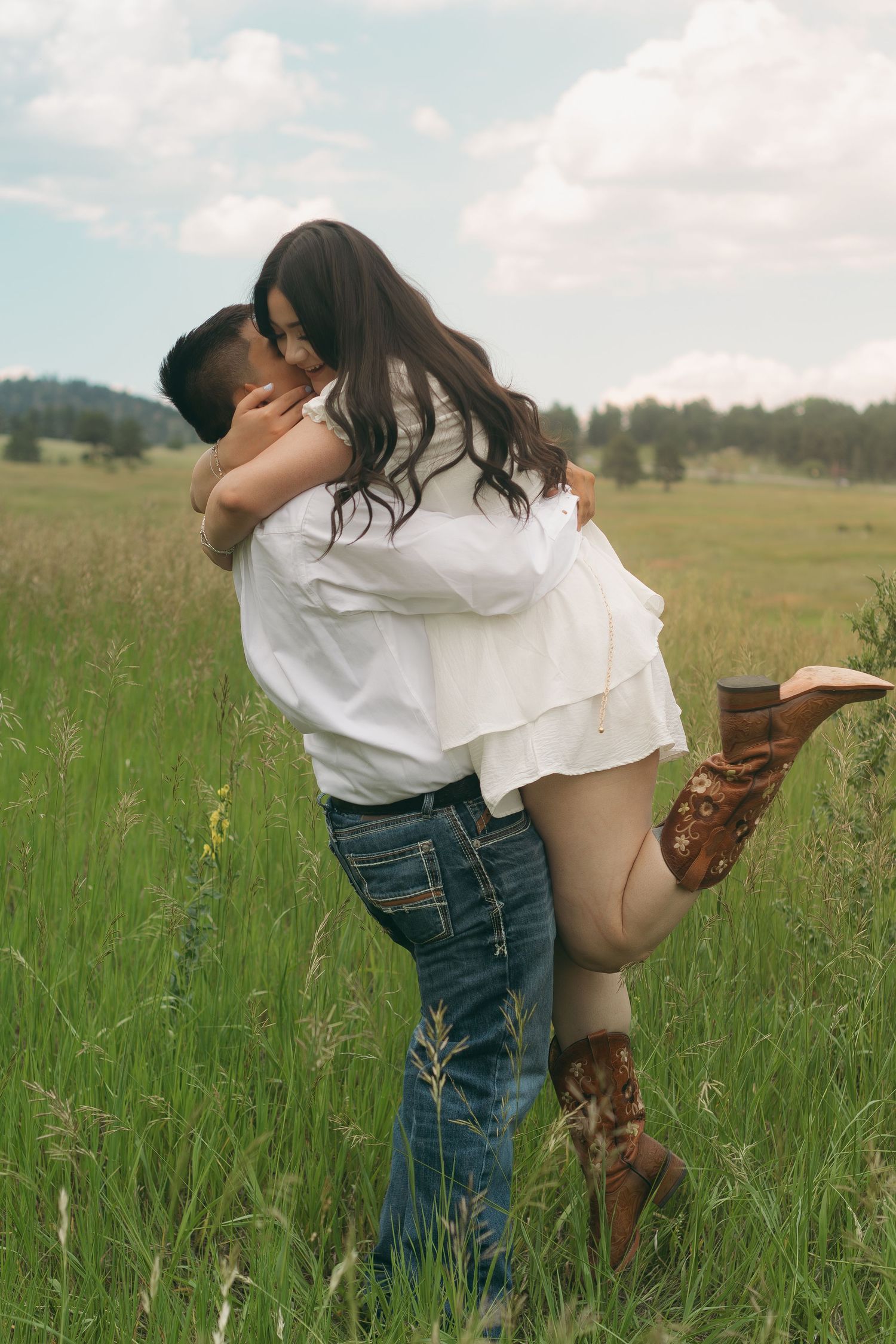 A couple shares intimate moments in a grassy field with mountains and cloudy skies in the background.