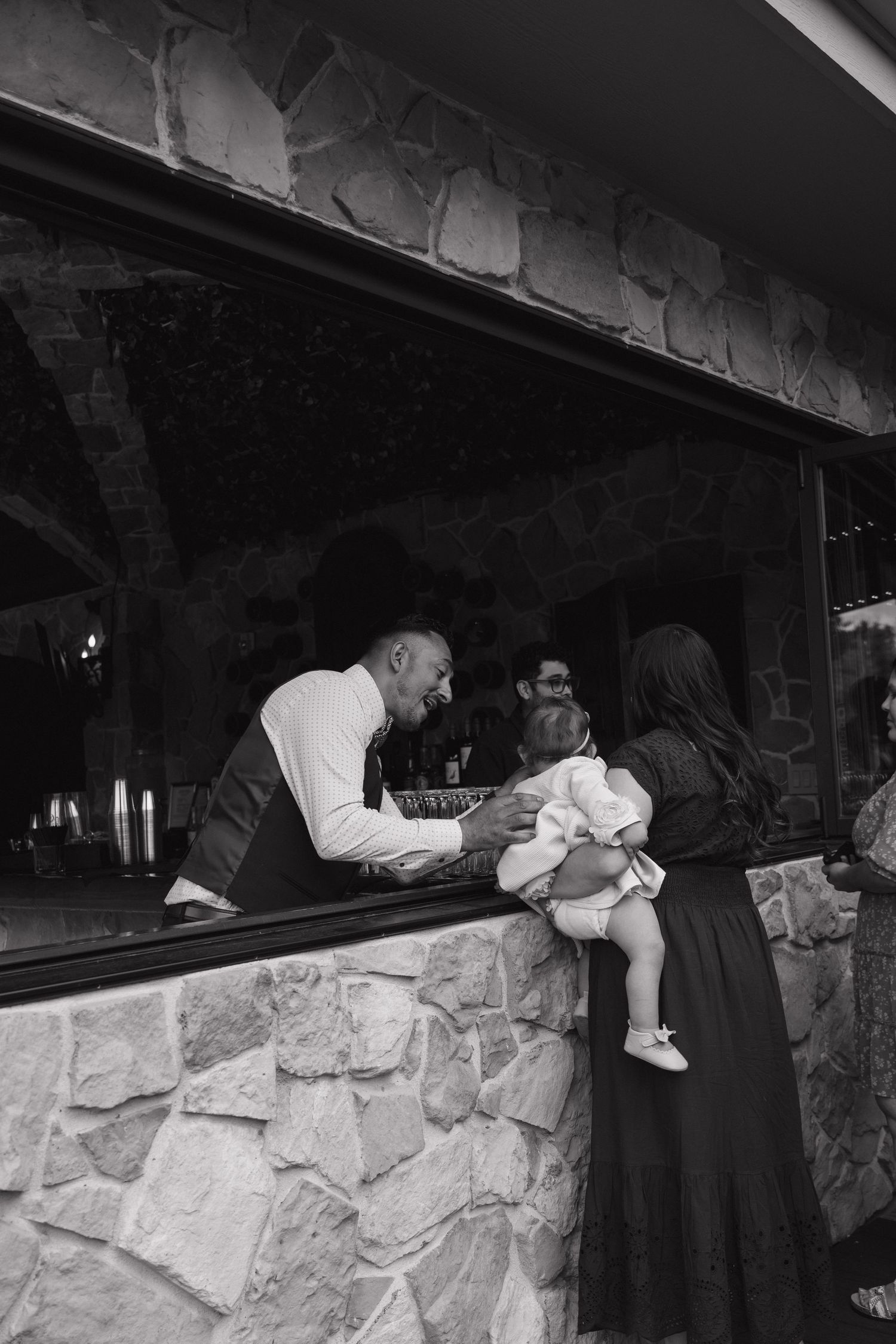 Black and white photo of a wedding guest holding a baby at a rustic stone bar during a wedding reception.