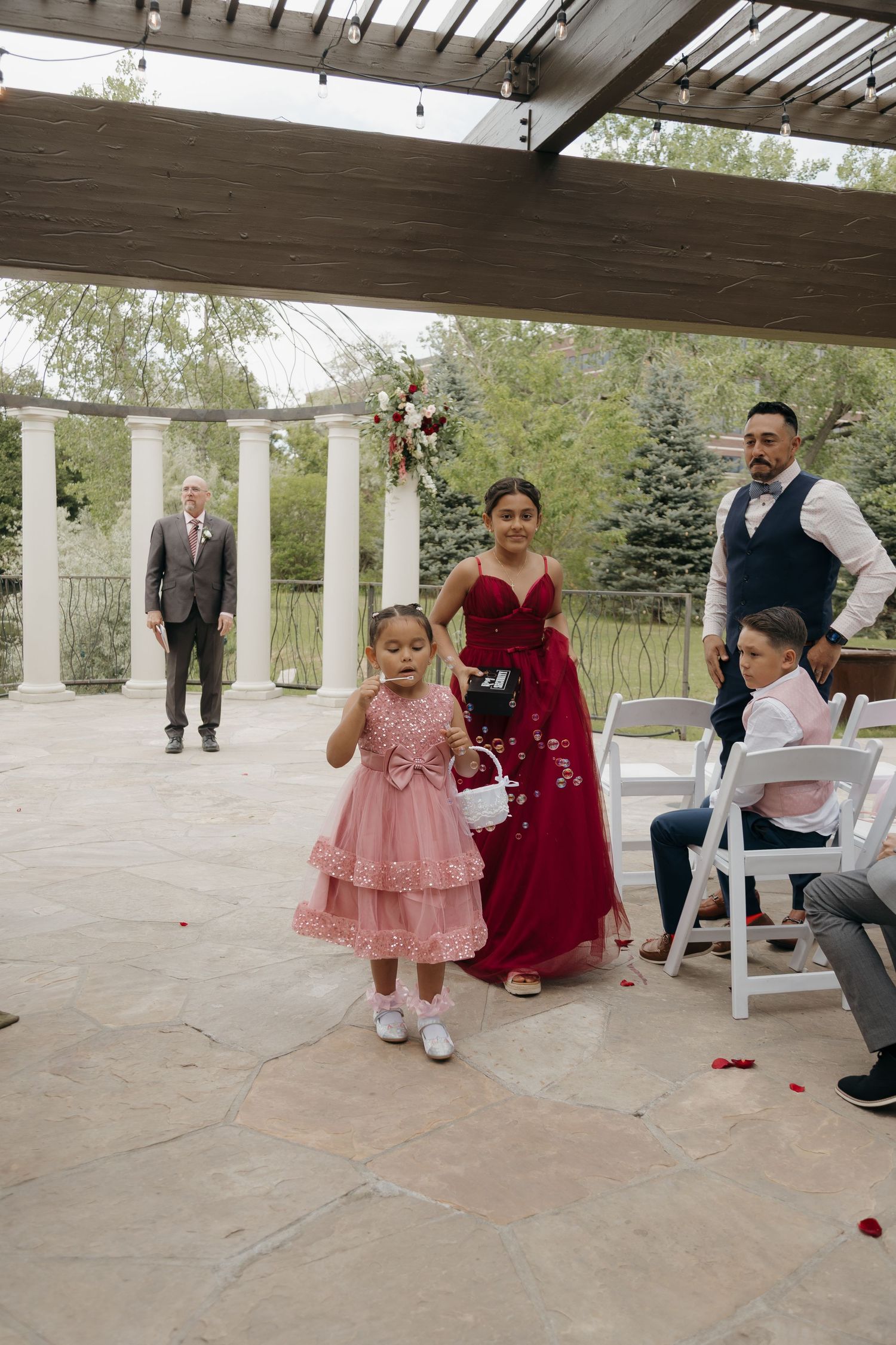 Young flower girl in pink dress scattering petals at outdoor wedding ceremony with white columns.