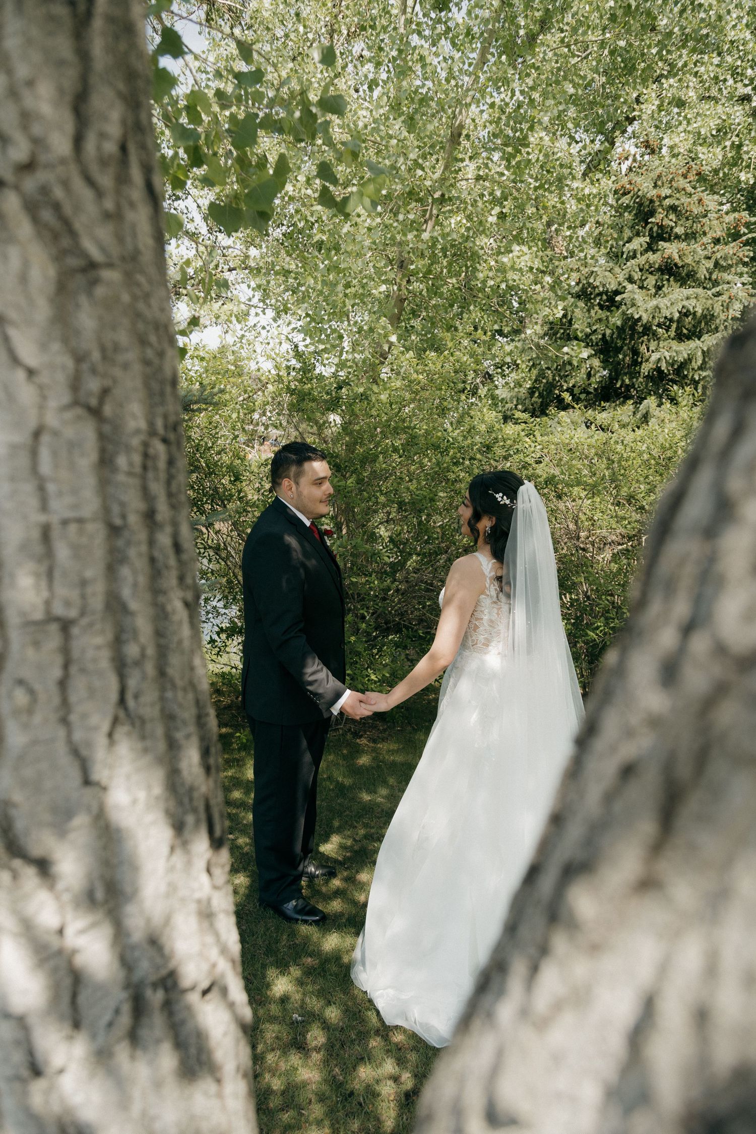 Bride and groom holding hands between two large trees in a lush green outdoor wedding setting.