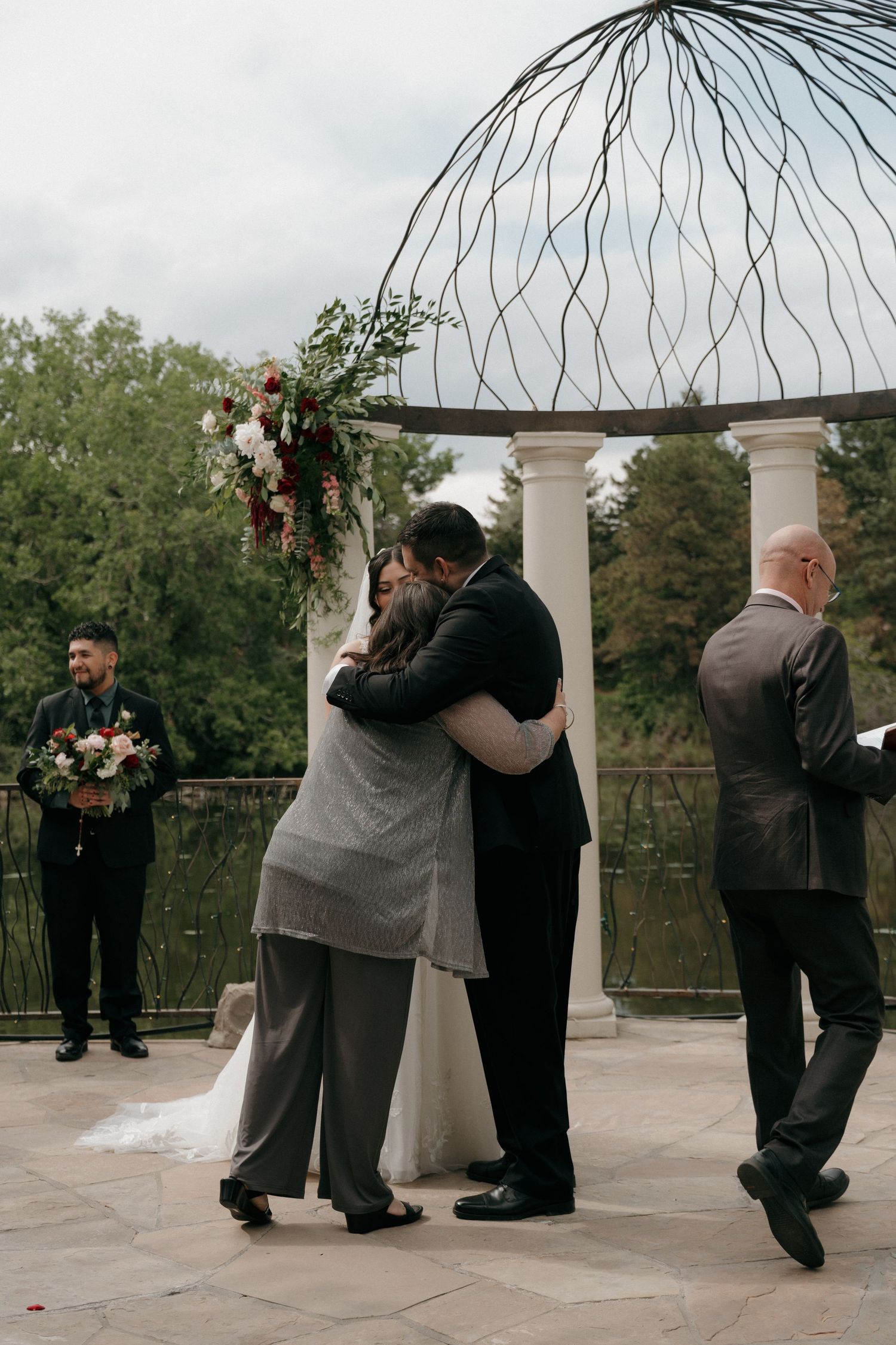 Bride and groom share emotional hug with family member at outdoor wedding altar.