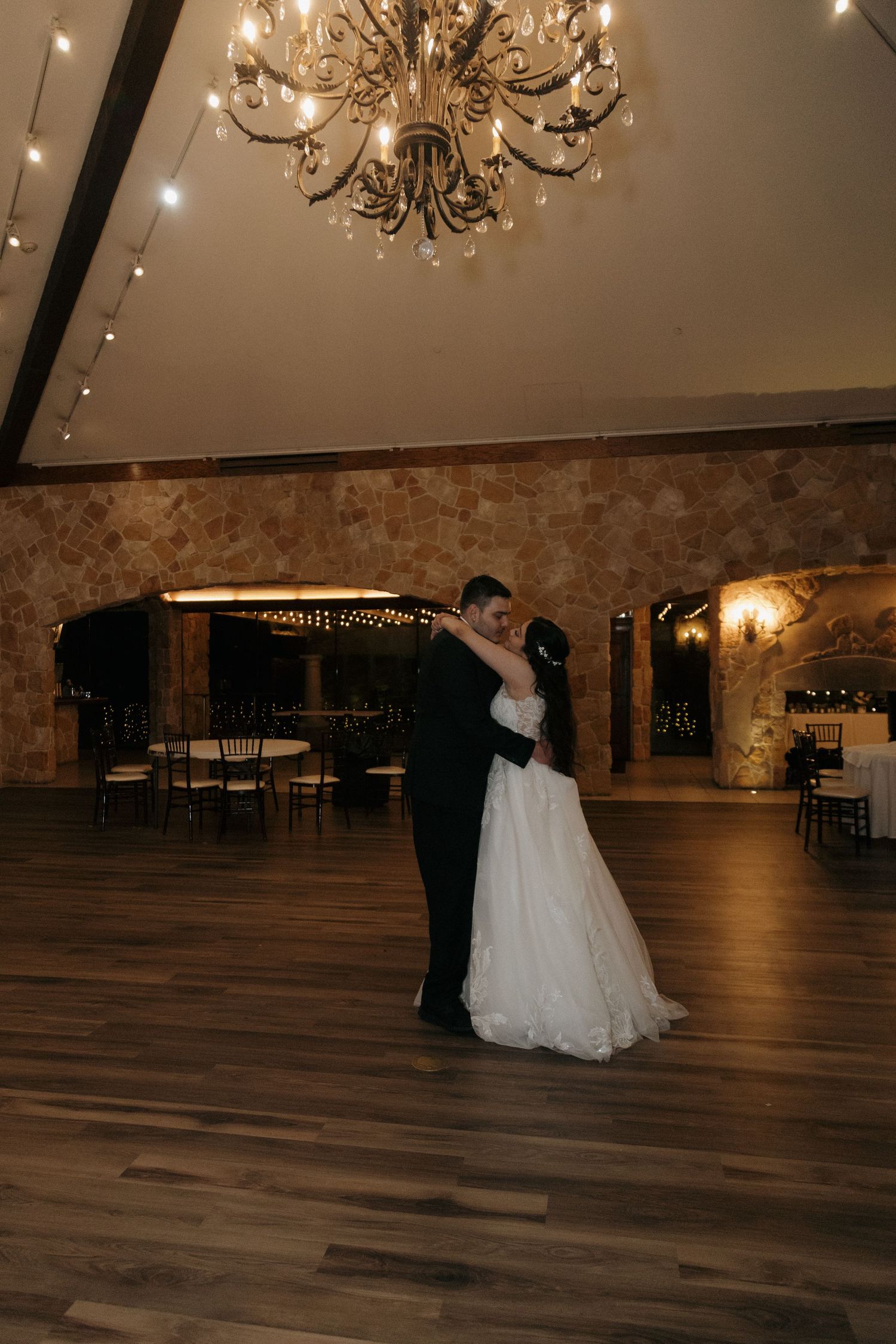 Bride and groom share their first dance under a glowing chandelier in an elegant rustic stone ballroom.