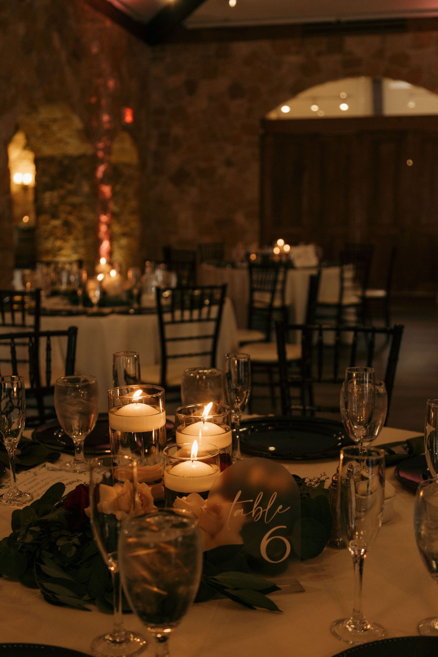 Romantic wedding reception table setting with floating candles, greenery, and a table number sign in a stone venue.