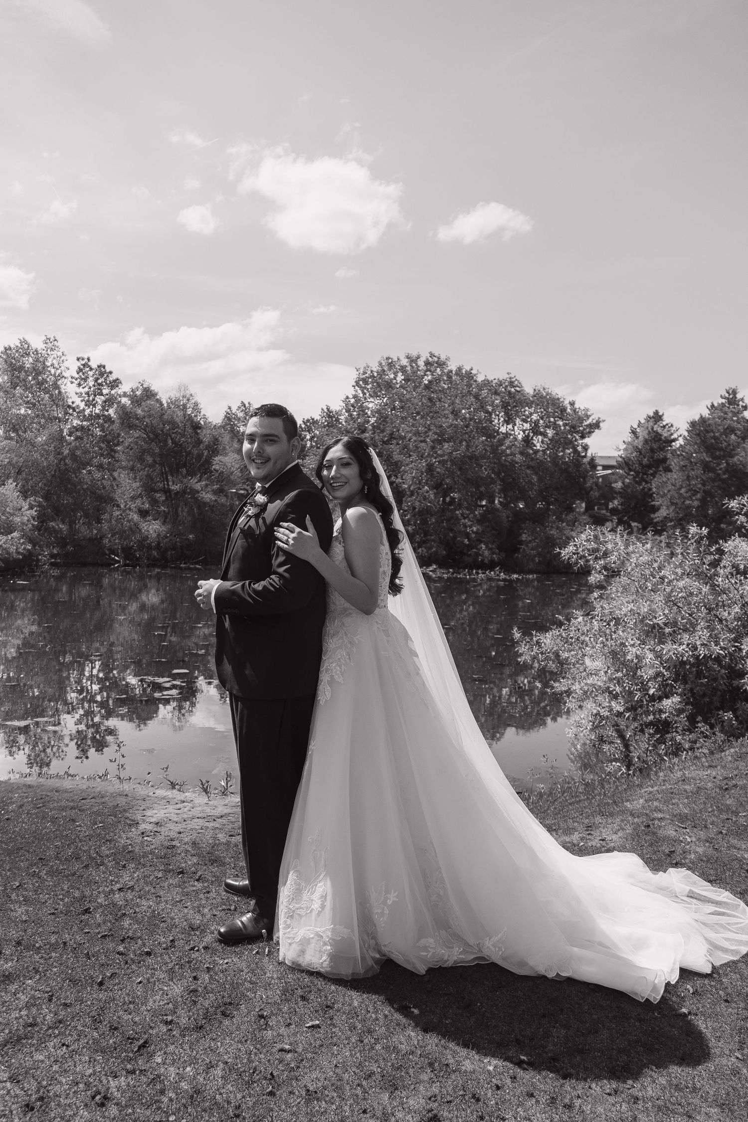 Black and white photo of bride and groom posing by a reflective lake surrounded by trees on their wedding day.
