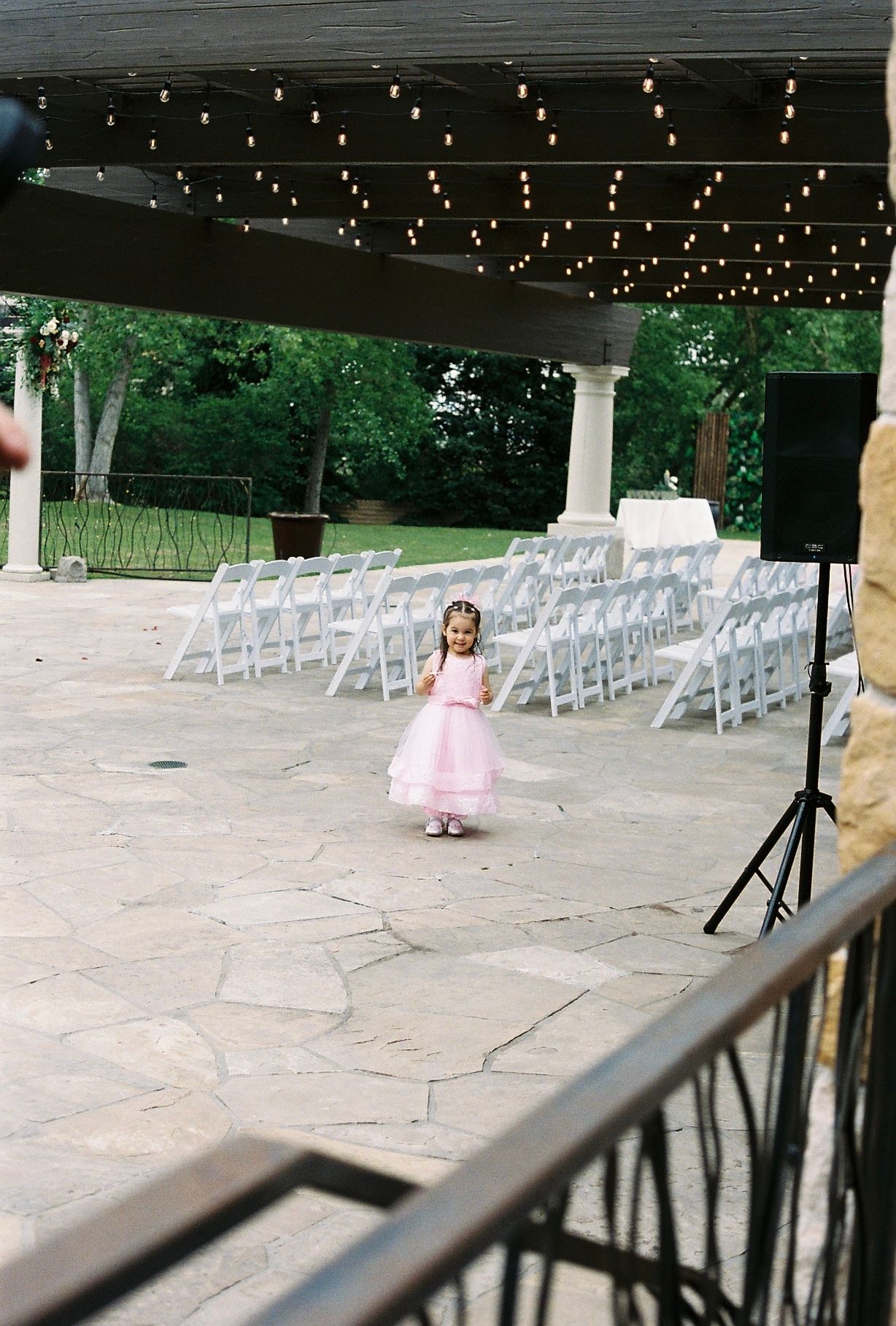 Young flower girl in pink dress standing alone on stone patio near rows of white chairs.