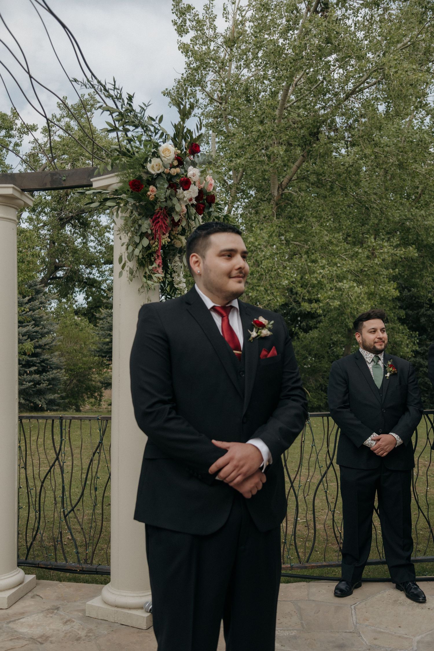 Groom in black suit with red tie stands at floral arch waiting at outdoor wedding ceremony.