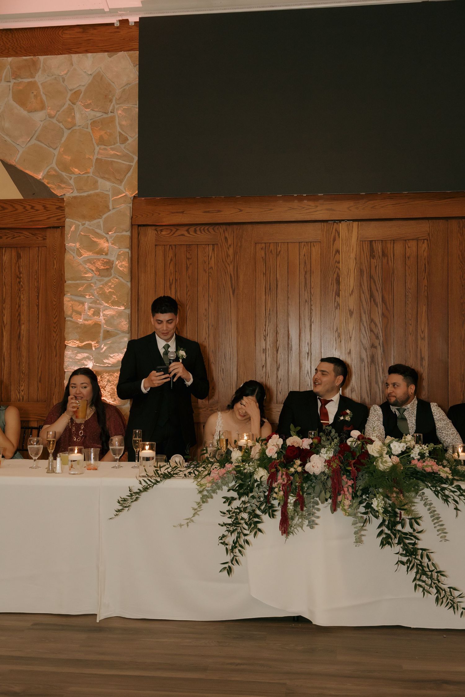 Best man delivering a wedding speech at the head table adorned with floral arrangements.