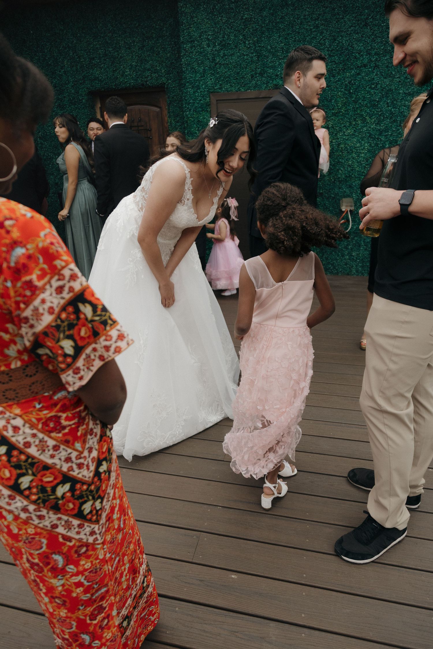 Bride in white lace gown bends down to dance with a young flower girl in pink at wedding reception.