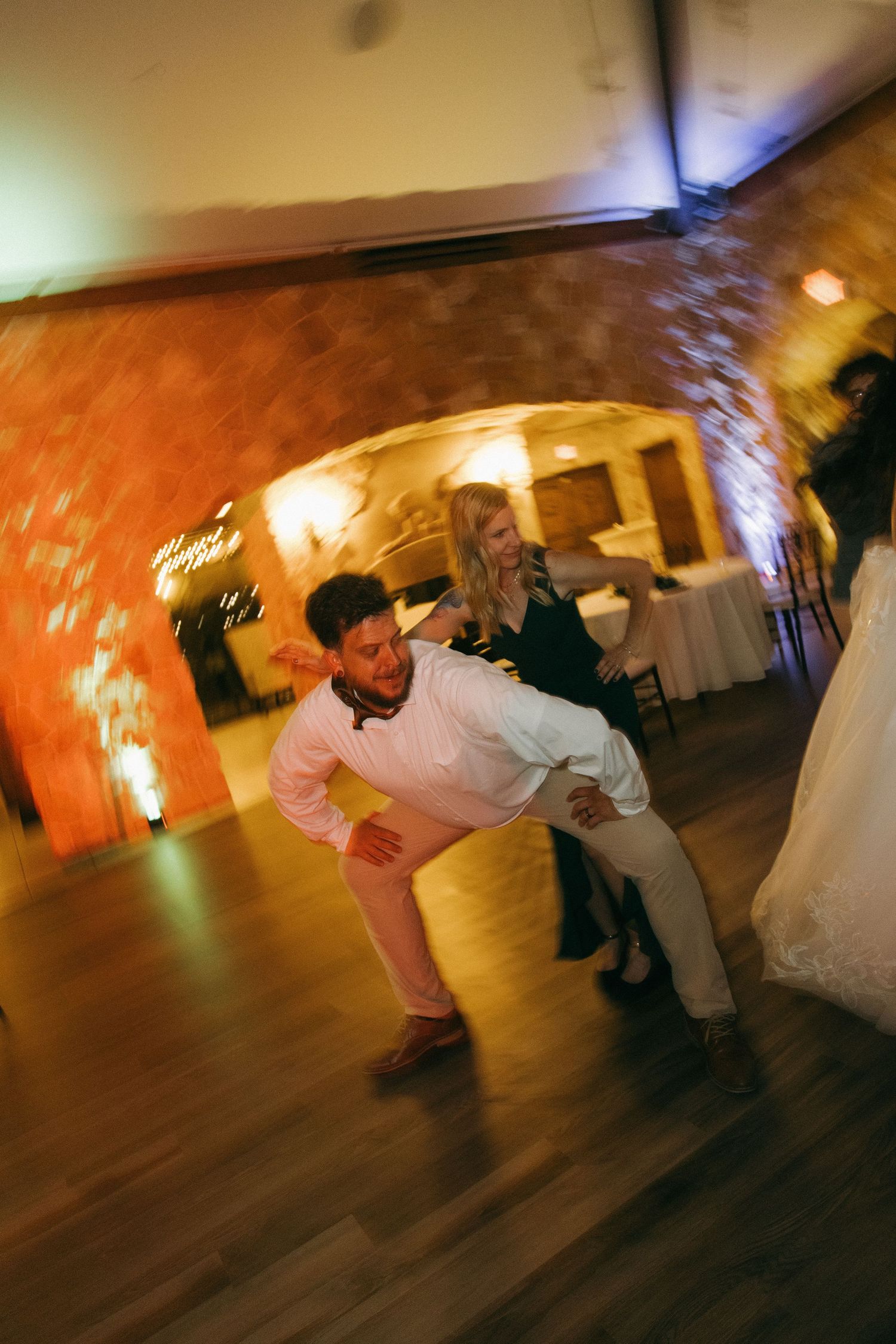 Energetic wedding guest dancing and leaning dramatically on the dance floor of a warmly lit stone venue.
