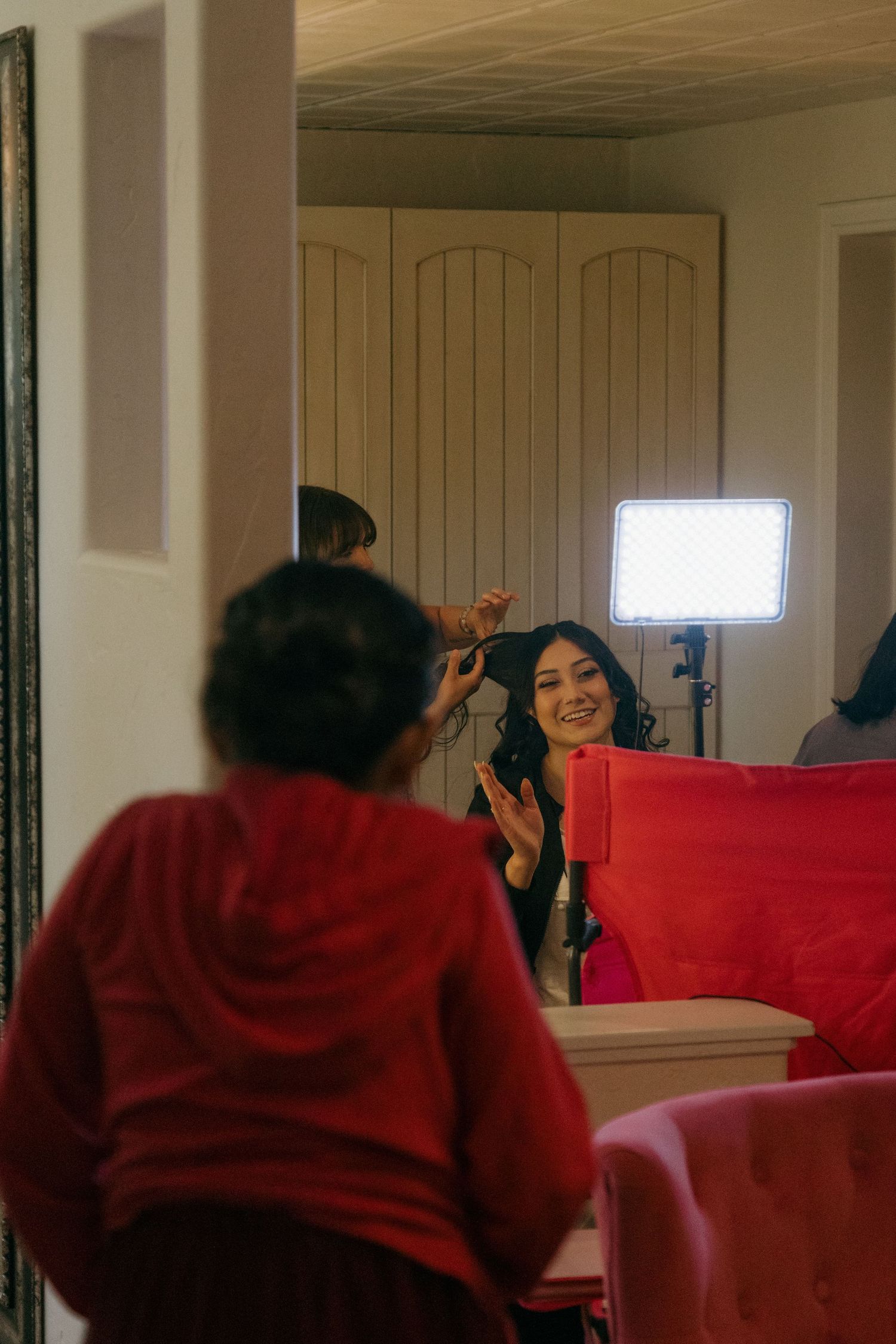Bride smiling at reflection in mirror during wedding hair and makeup preparation with bright studio lighting.