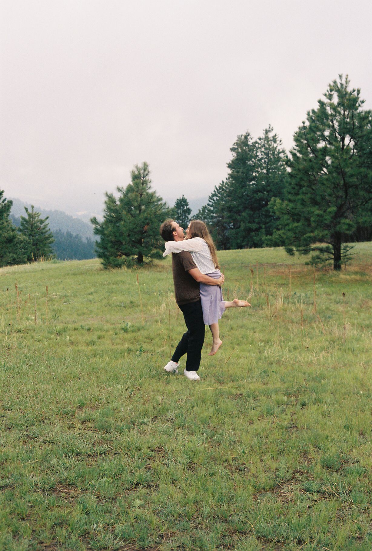 Man lifting woman in an embrace on a green meadow with pine trees and misty mountains behind.