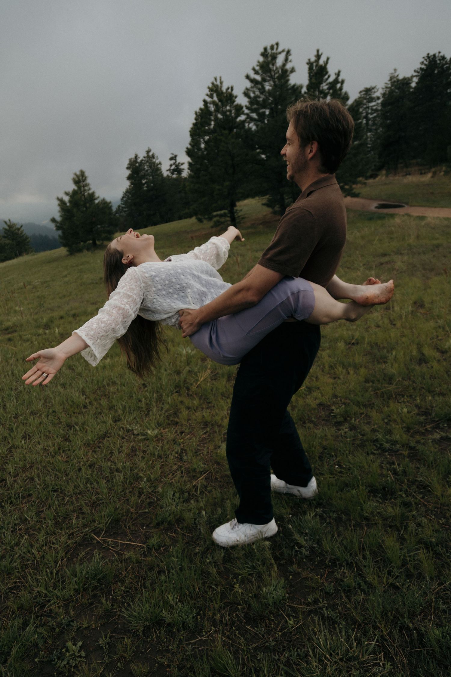 Man lifts smiling woman in his arms on a green meadow with pine trees in the background on a cloudy day.
