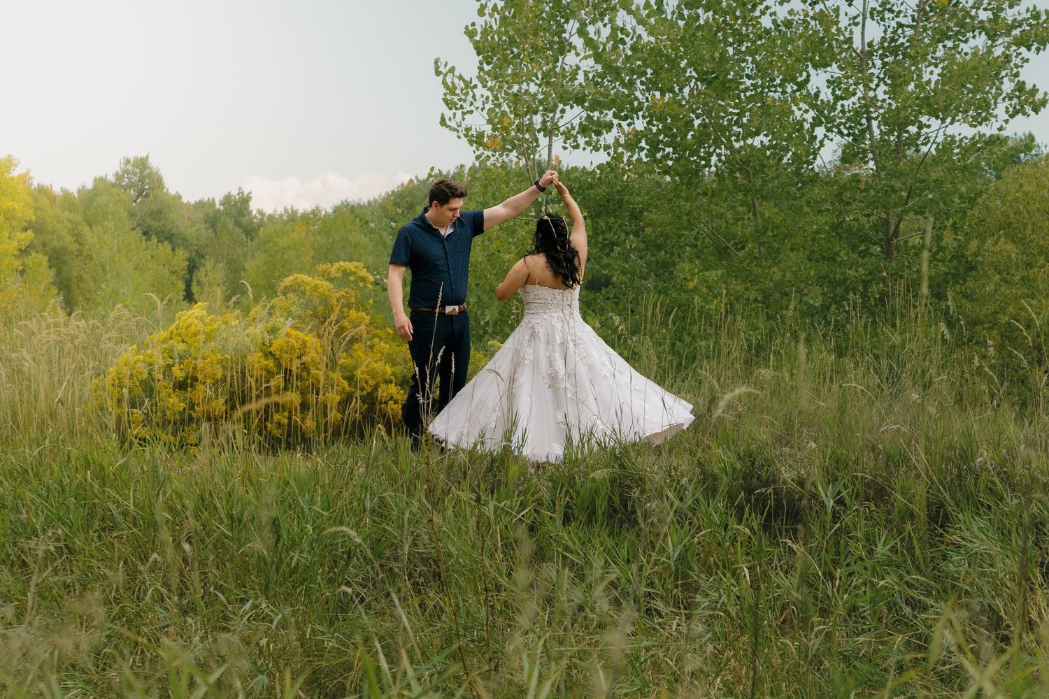 Romantic couple twirls in golden sunlight against a natural outdoor backdrop of trees and grass.