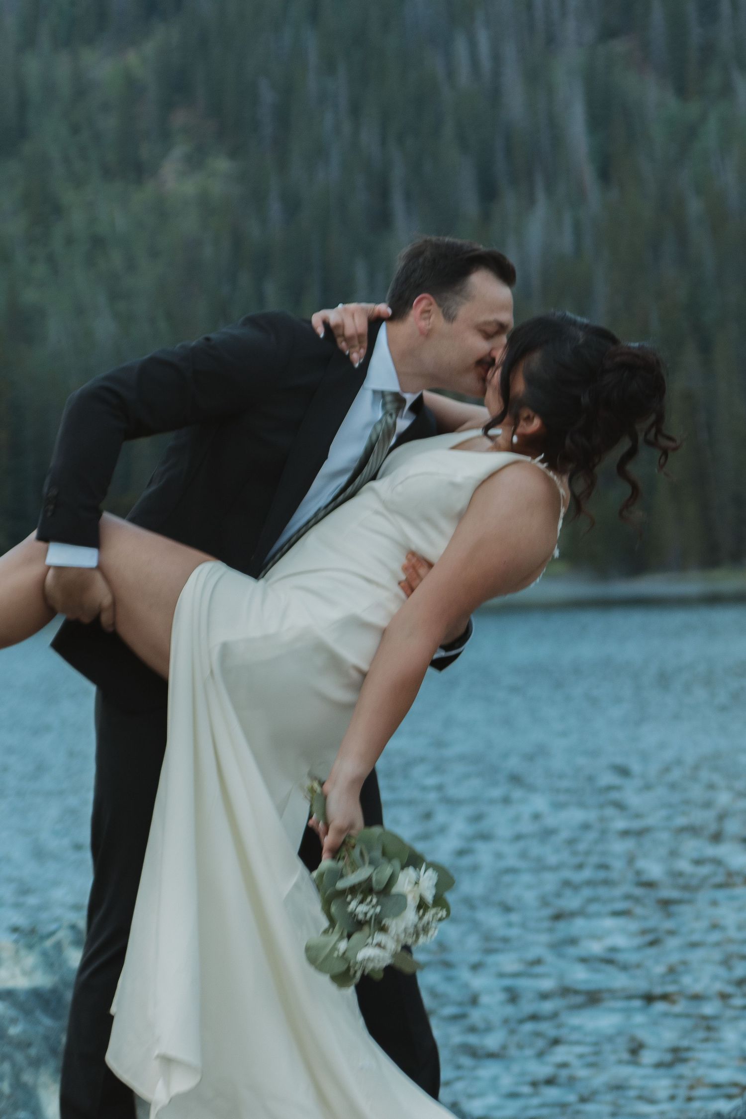 A romantic wedding couple embraces in a dramatic dip pose against a mountain lake backdrop.