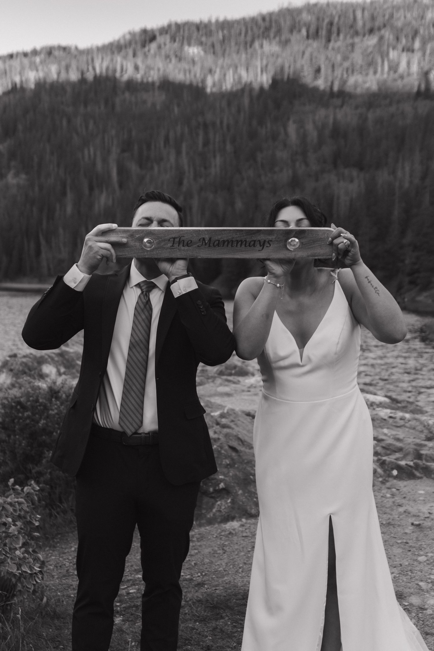 A wedding couple poses playfully near a scenic mountain lake lifting a bar weight together in formal attire.