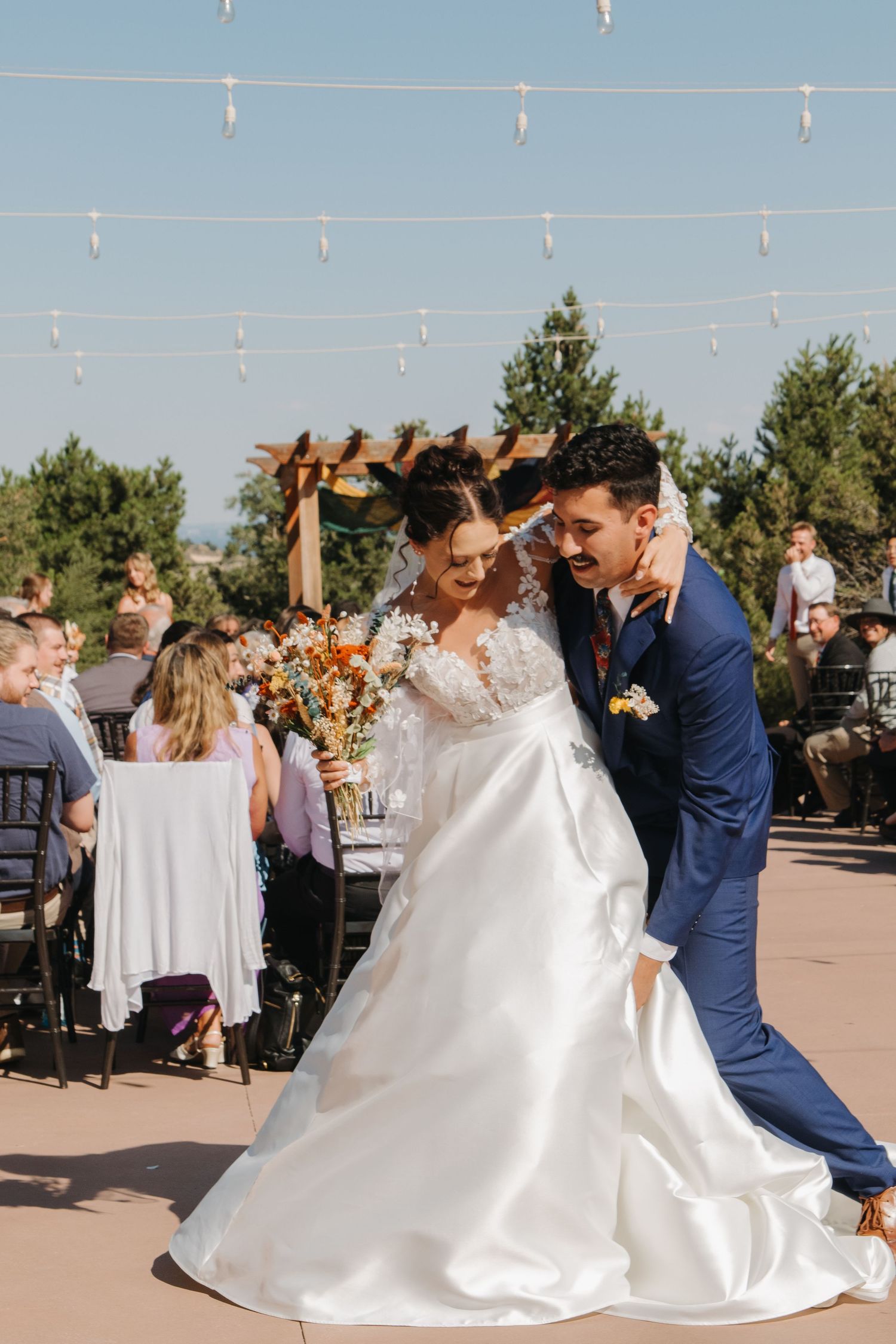 Couple performs first dance at outdoor wedding reception under string lights and blue sky.