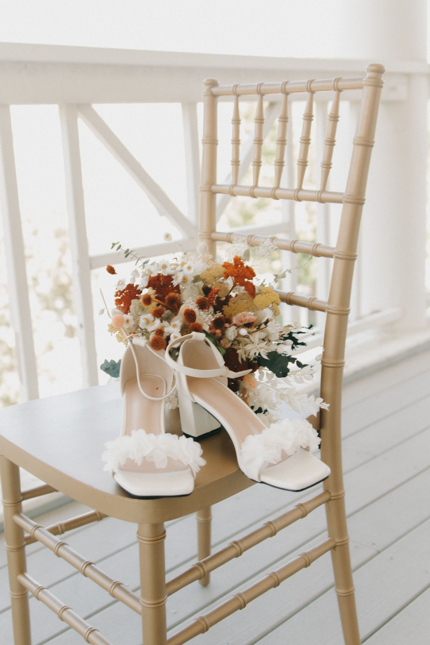 White bridal shoes adorned with flowers displayed on light wooden Chiavari chairs.
