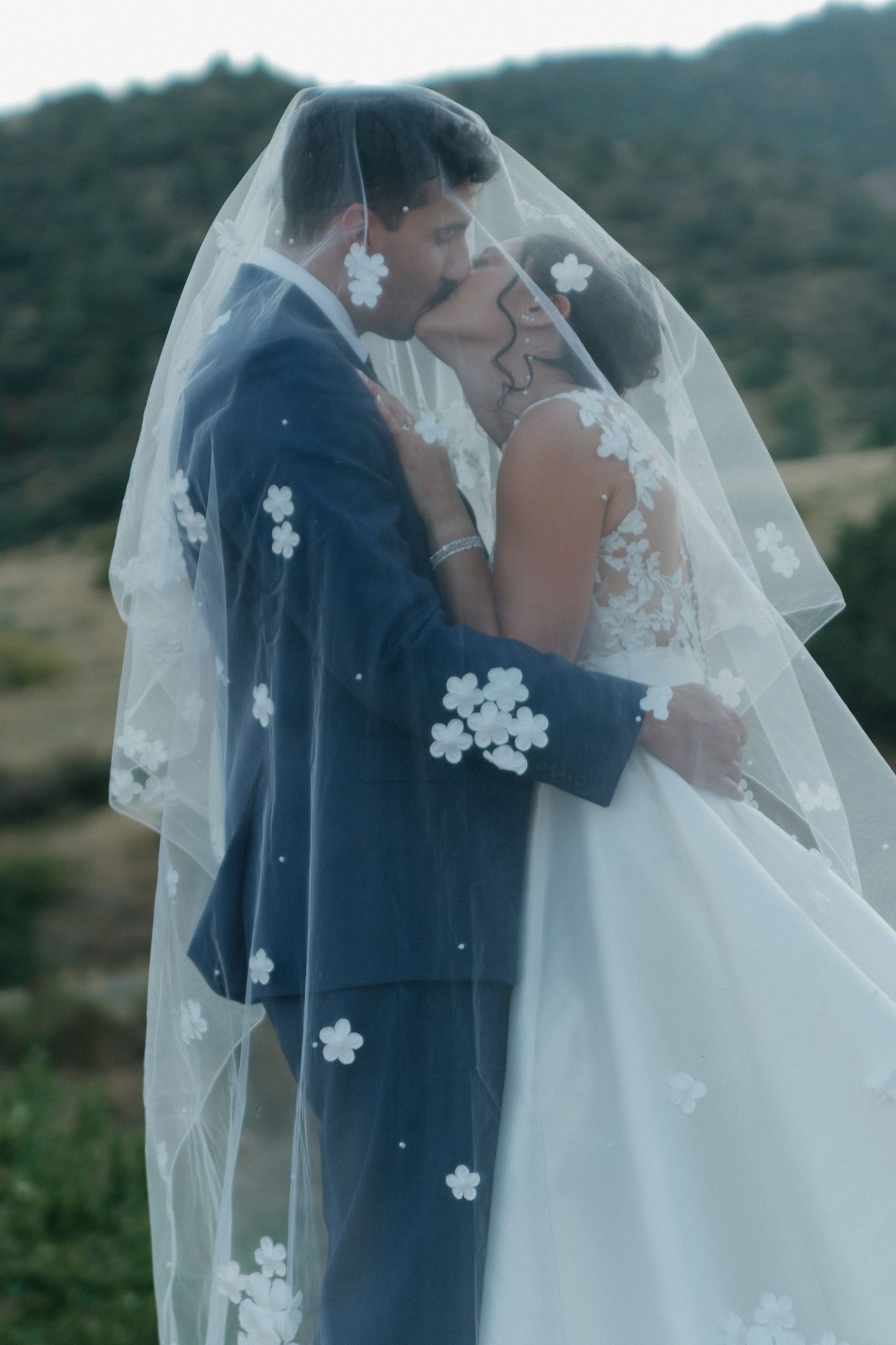 Romantic moment under floral embellished veil with mountain scenery in background.