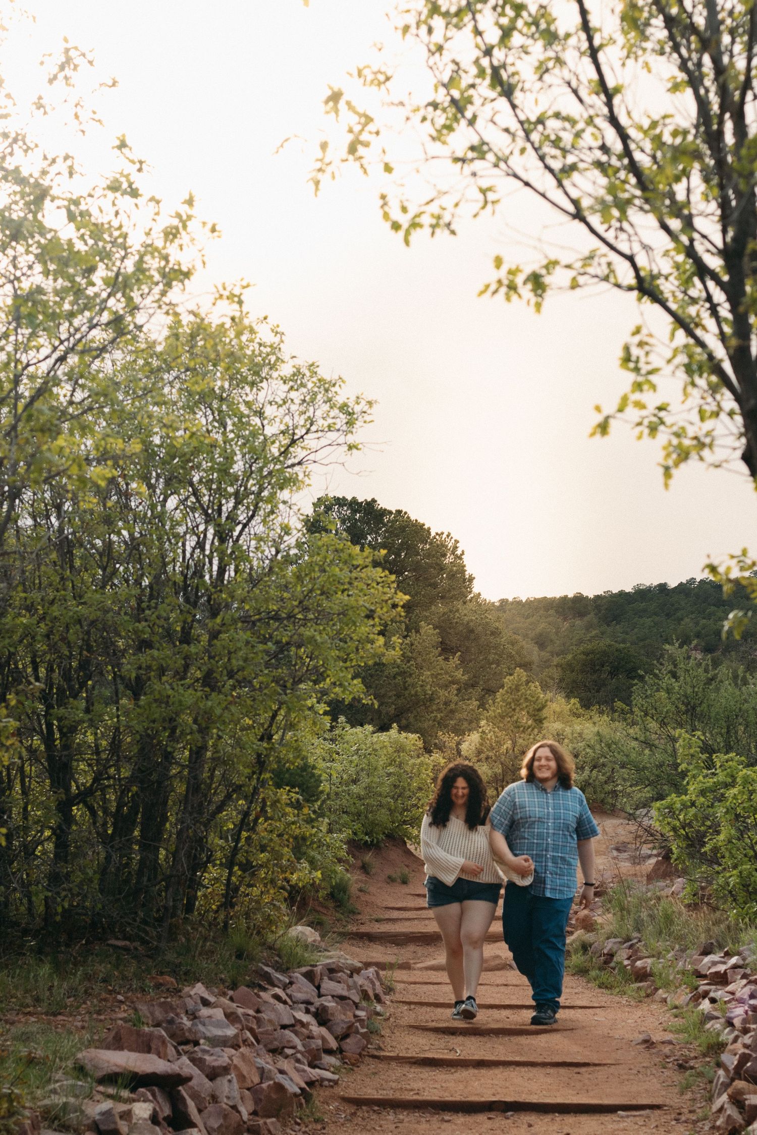 Two people walking hand-in-hand on a scenic hiking trail surrounded by green trees at sunset.