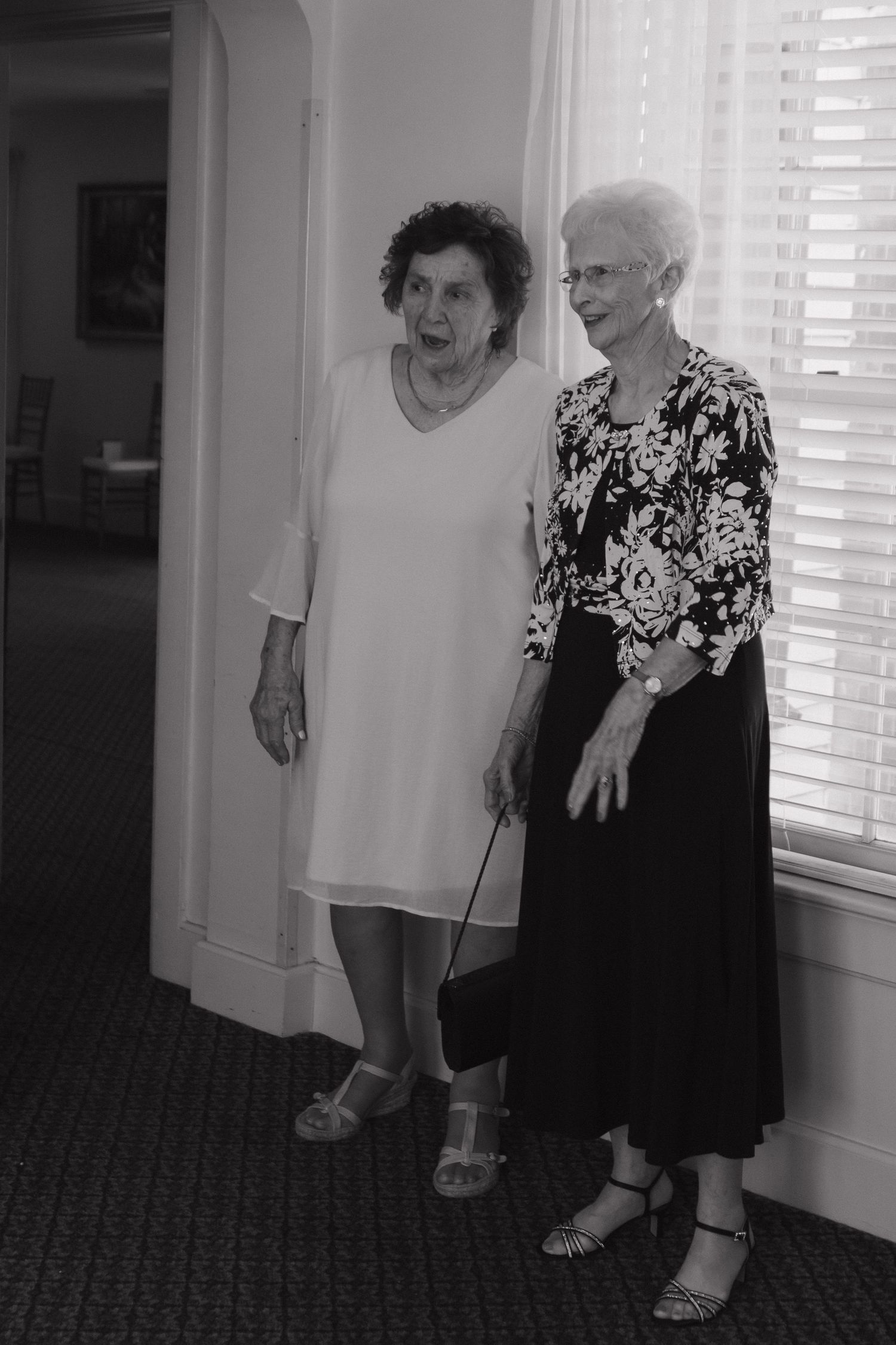 Two elderly people in casual attire laughing together in a hallway with white walls and carpeted floors.