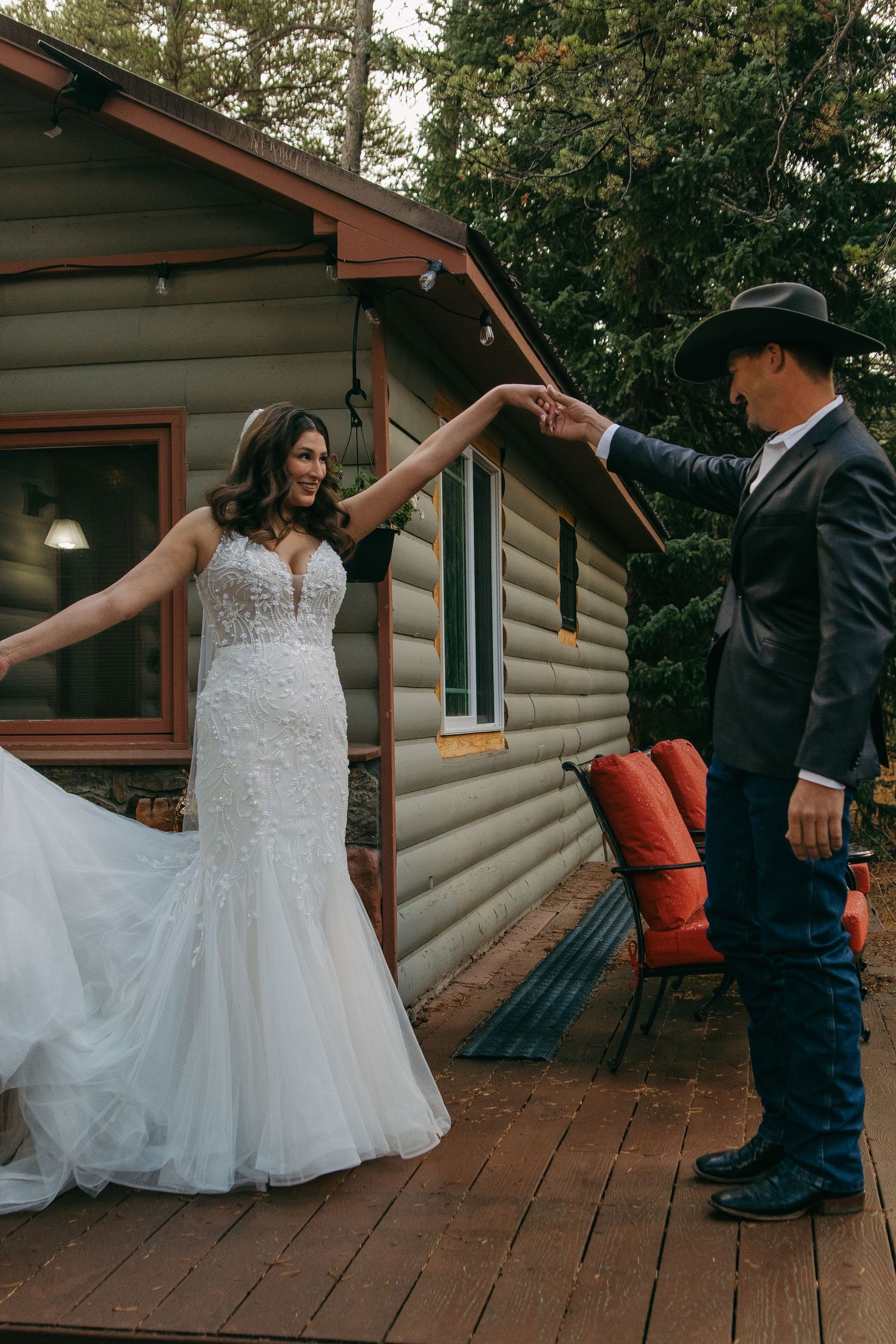A couple dances on a rustic wooden deck in front of a log cabin, with the bride wearing a flowing white gown.