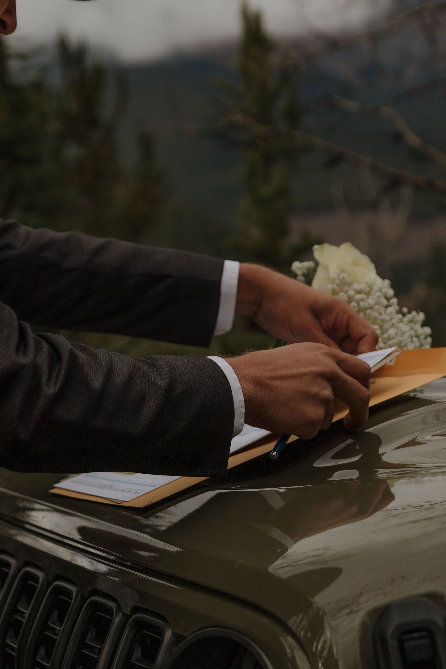 Hands holding wedding vows on a car hood with mountain scenery in the background.