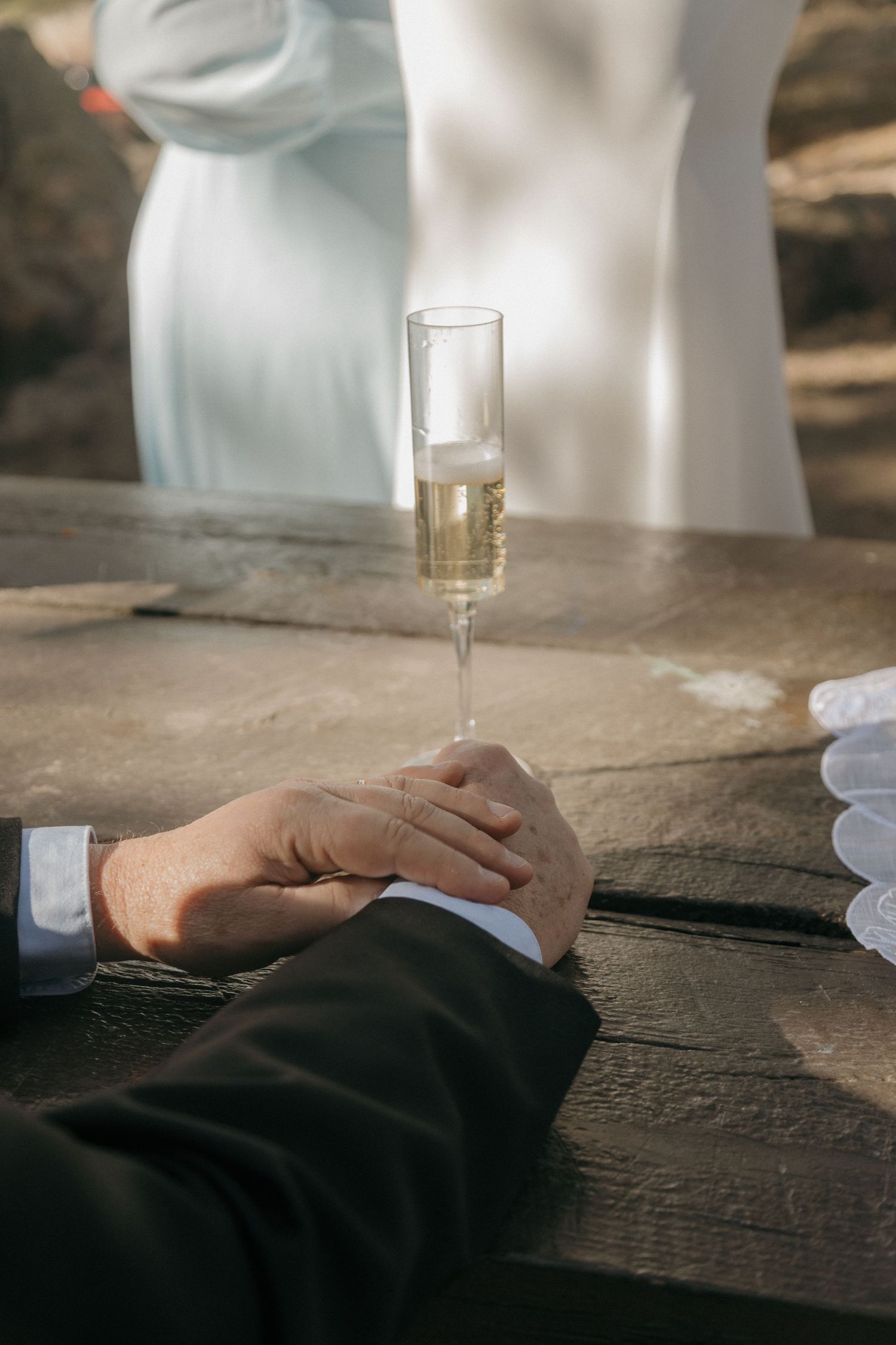 Close-up of couple holding hands beside a champagne flute on a rustic wooden surface during wedding celebration.