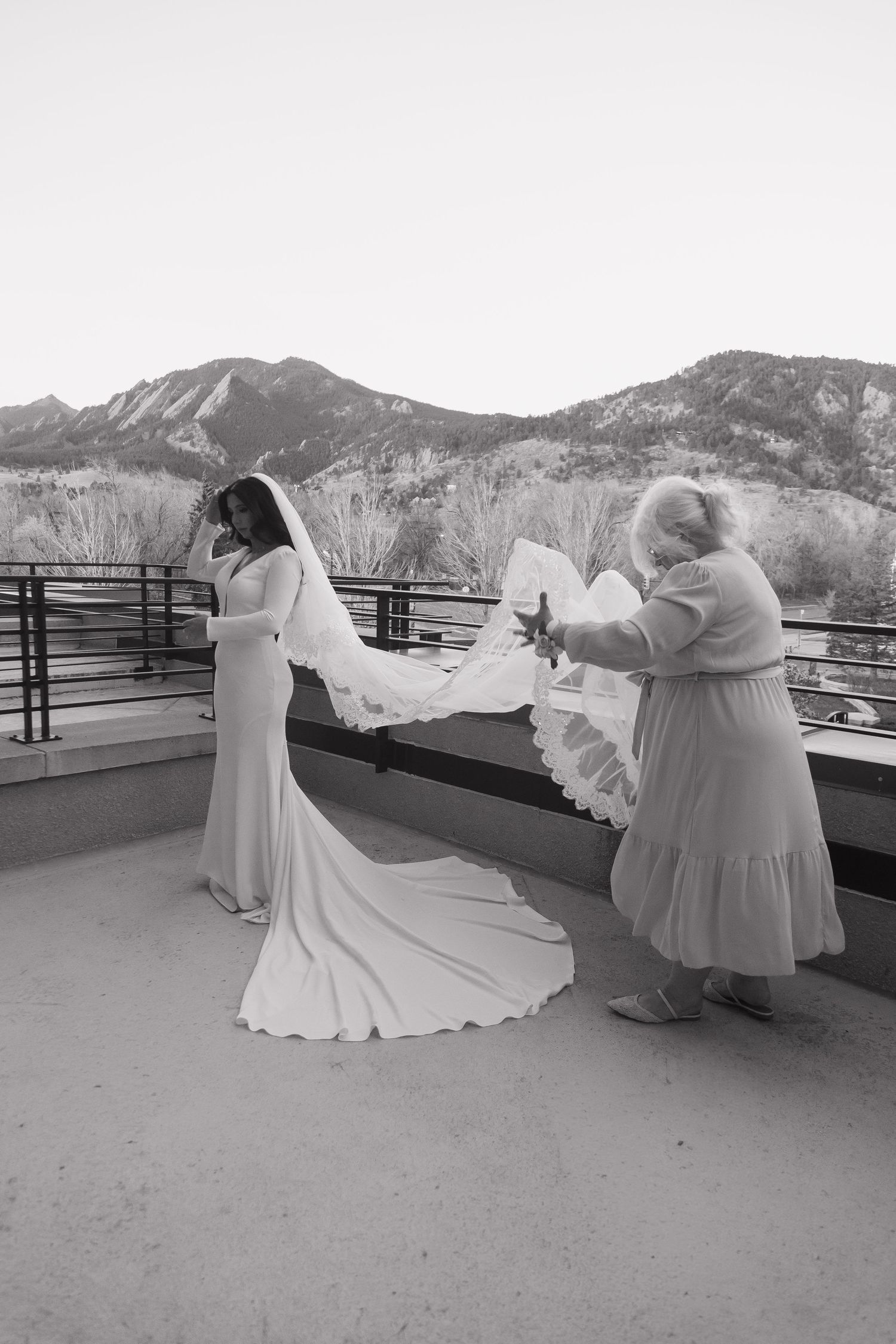 Black and white photo of bride in elegant gown with long veil being fluffed by assistant on mountain overlook.