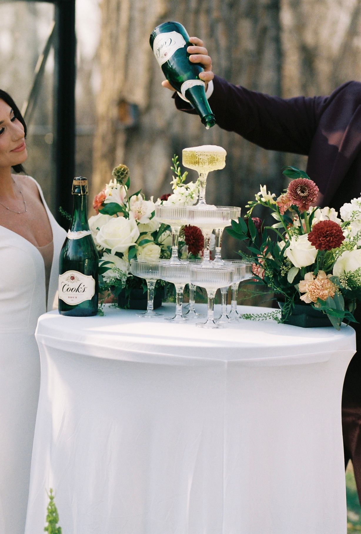 Groom pours champagne into a tower of glasses beside bride in white dress at a floral-decorated wedding reception table.