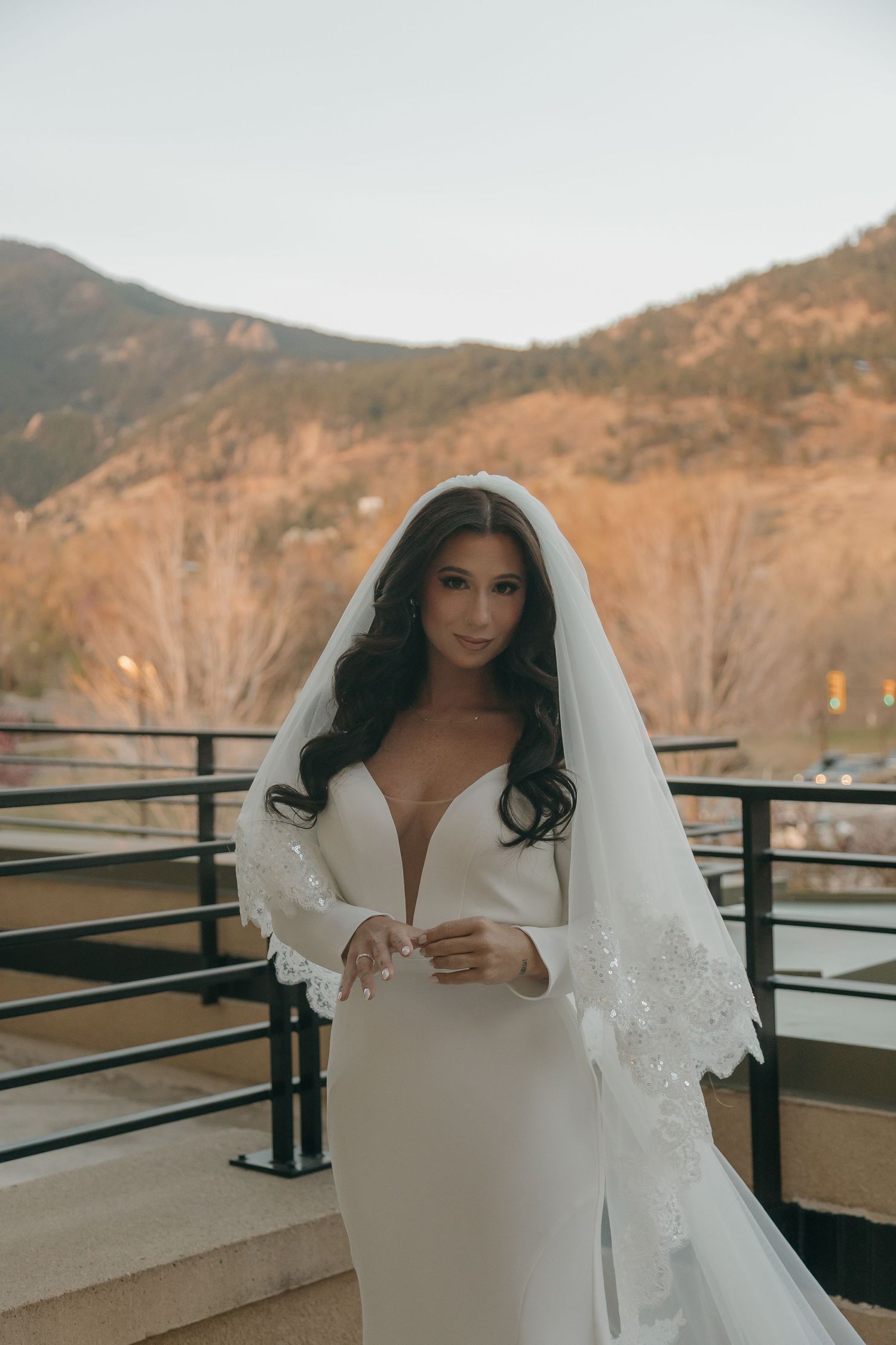 An elegant bride in a deep V-neck gown and lace-trimmed veil poses on a mountain balcony.
