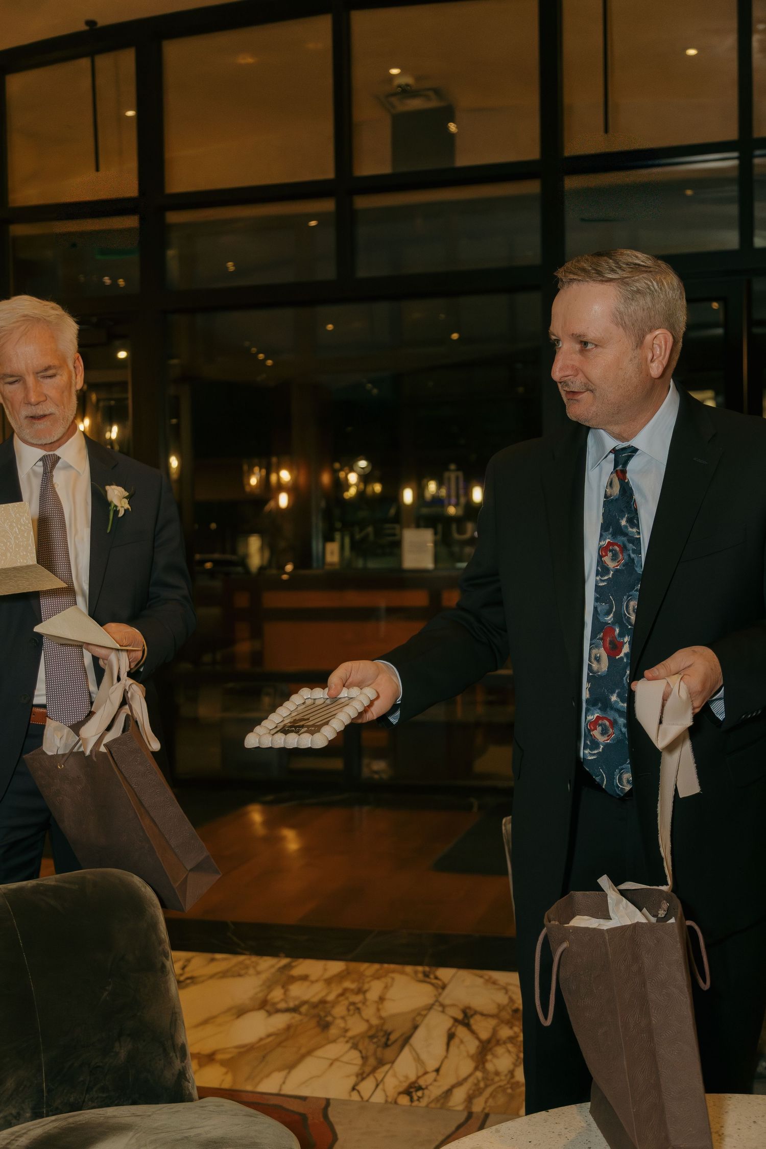 Two men in suits exchange wedding gifts in an elegant hotel lobby with black-framed glass windows.