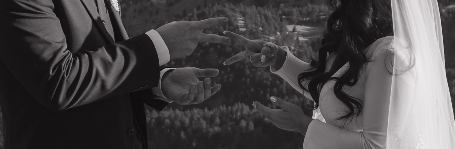 Black and white close-up of bride and groom exchanging rings during intimate outdoor mountain wedding ceremony.