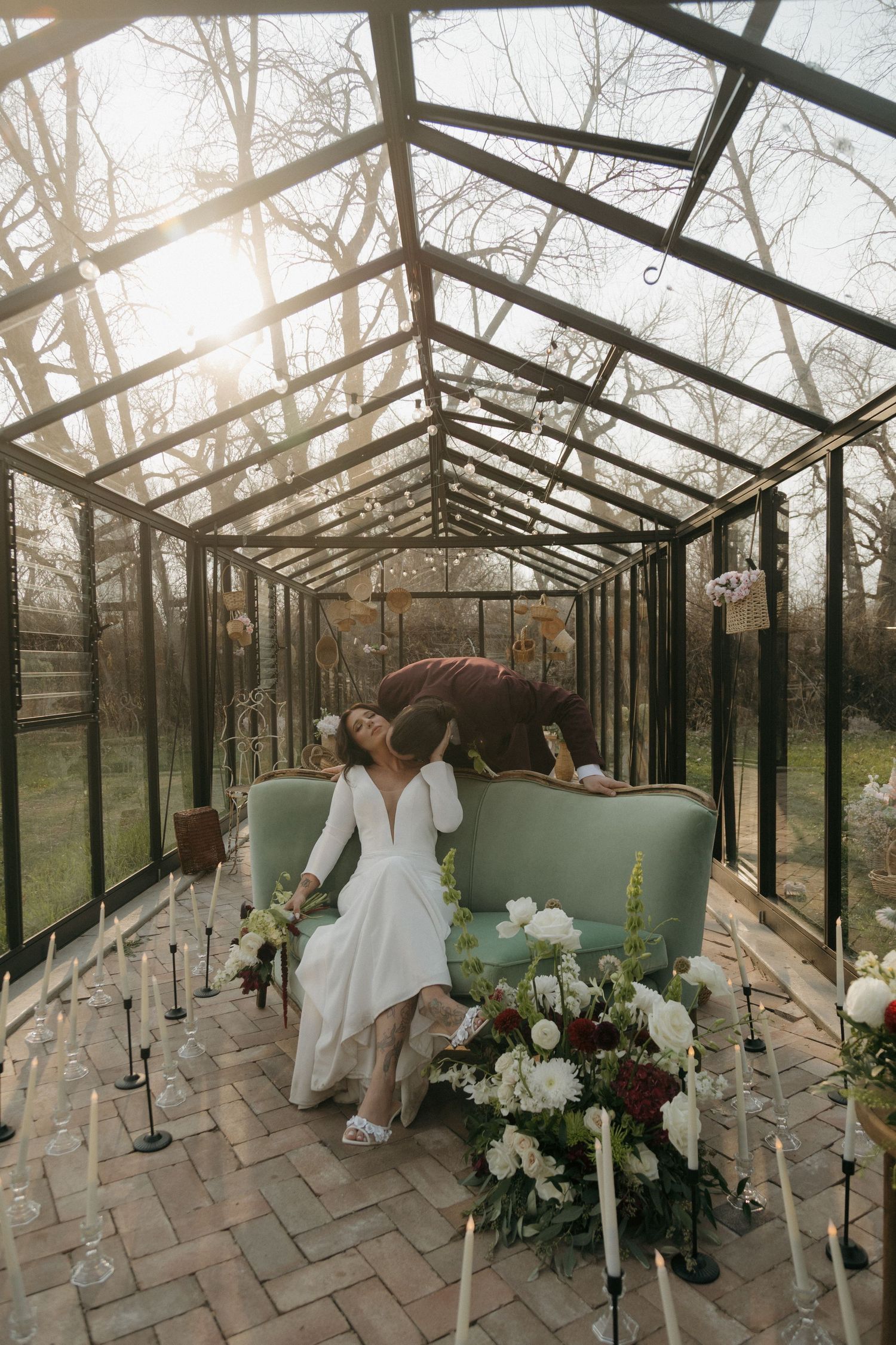 Bride in white dress sits on a mint sofa inside a sunlit glass greenhouse decorated with candles.