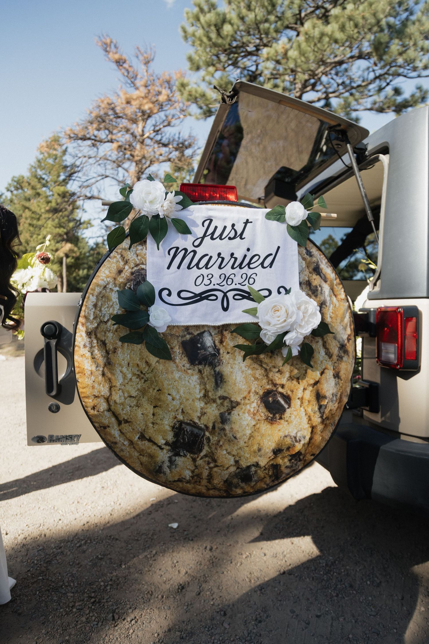 Jeep spare tire cover decorated with Just Married 03.26.26 sign and white roses at outdoor wedding.