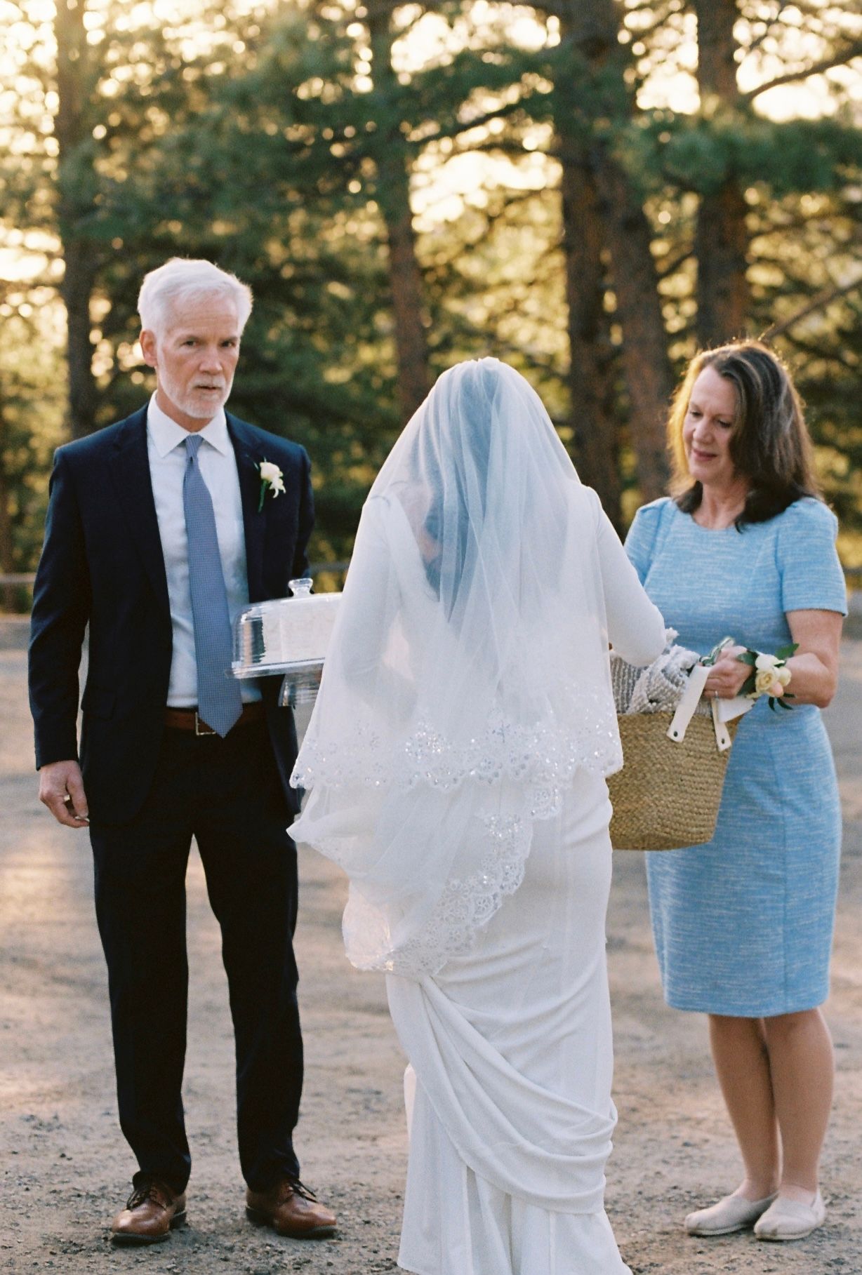Veiled bride walking with parents along forest path during golden hour outdoor wedding ceremony.