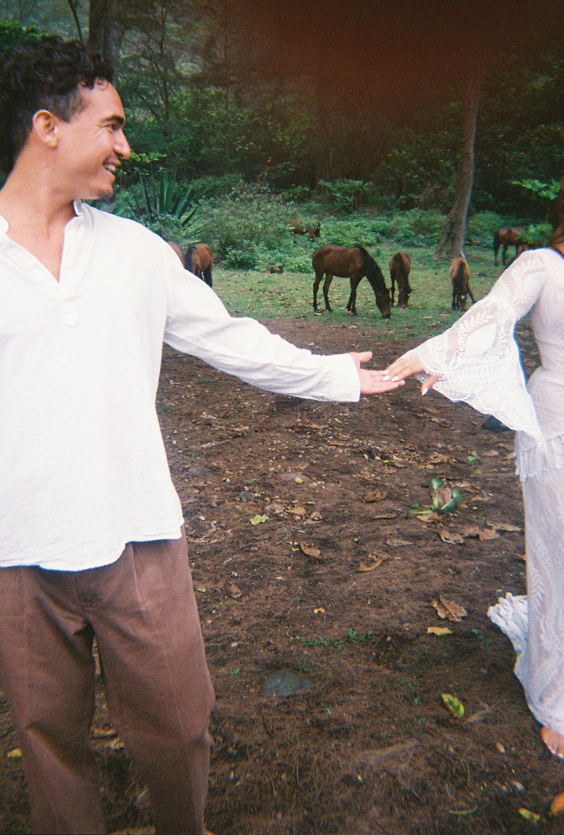Groom in white shirt smiling while holding bride's hand in lace dress with grazing horses behind them.