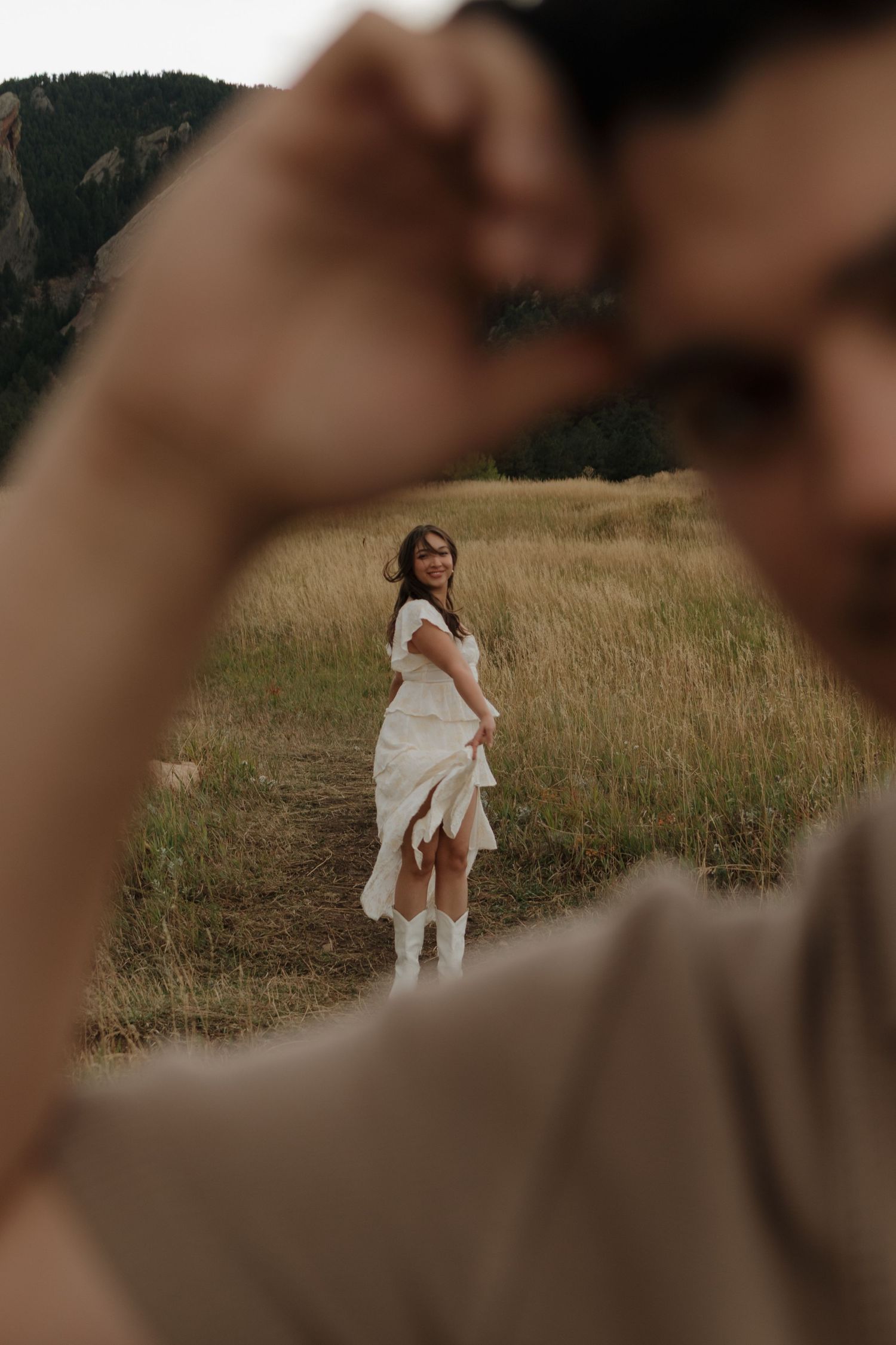 Bride in white ruffled dress and cowboy boots smiles in mountain meadow, framed by blurred foreground figure.