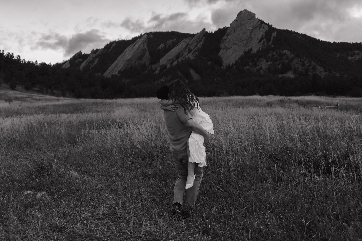 Couple embracing in a grassy field with dramatic rocky Flatirons mountains in black and white.