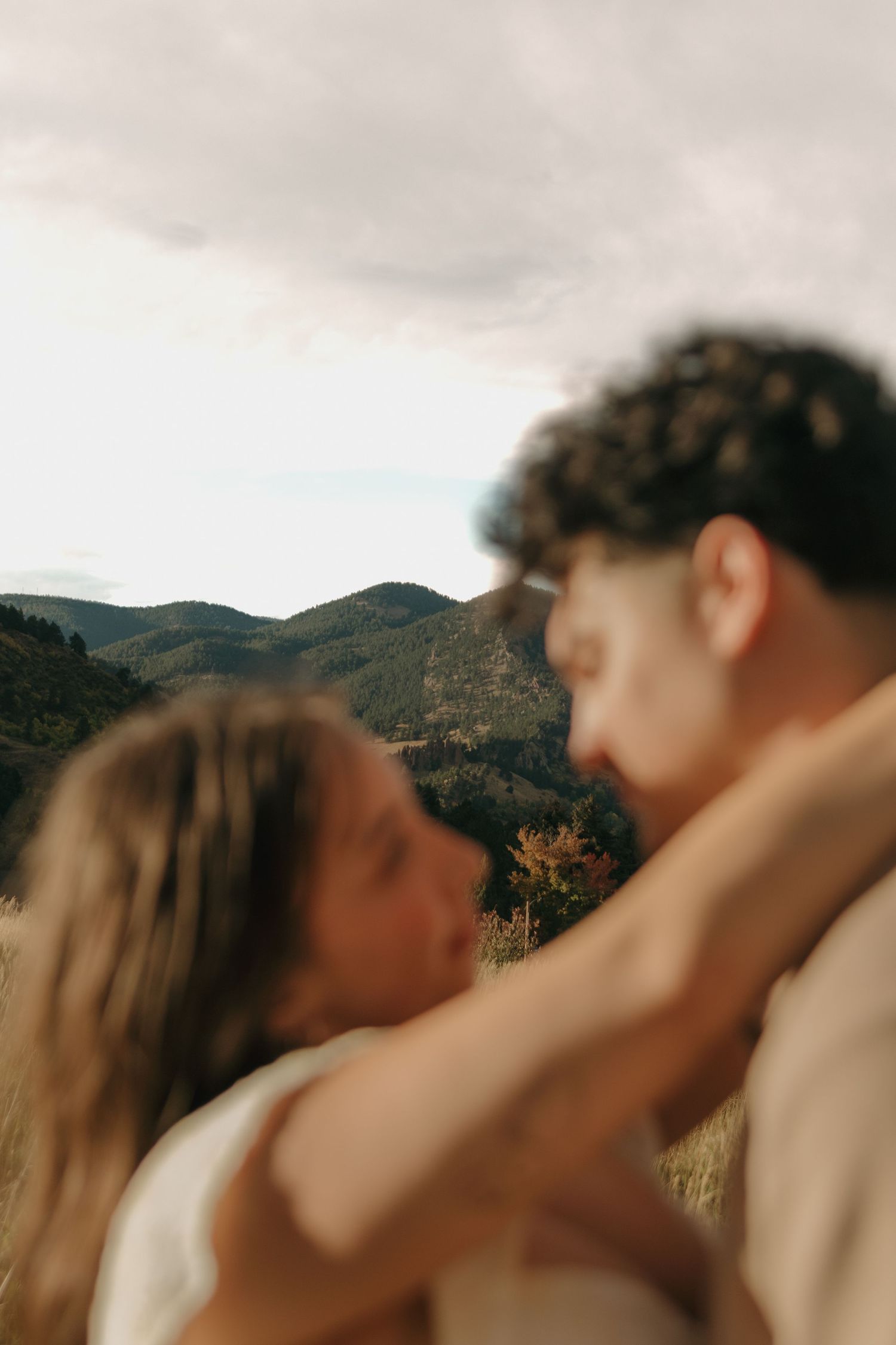Blurred romantic couple embracing with scenic mountain landscape and cloudy sky in background.