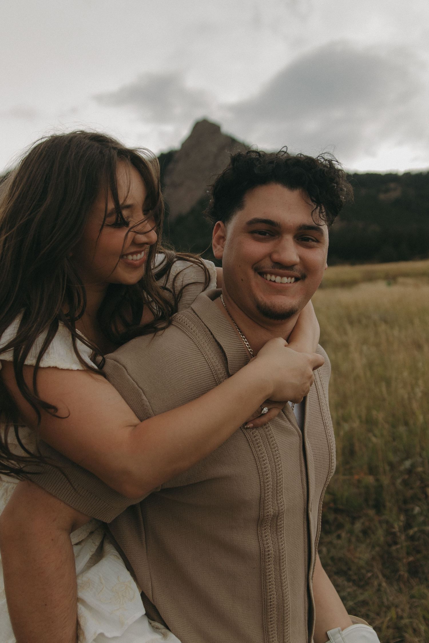 A joyful couple laughing together in a field, the woman hugging the man from behind near rocky mountains.