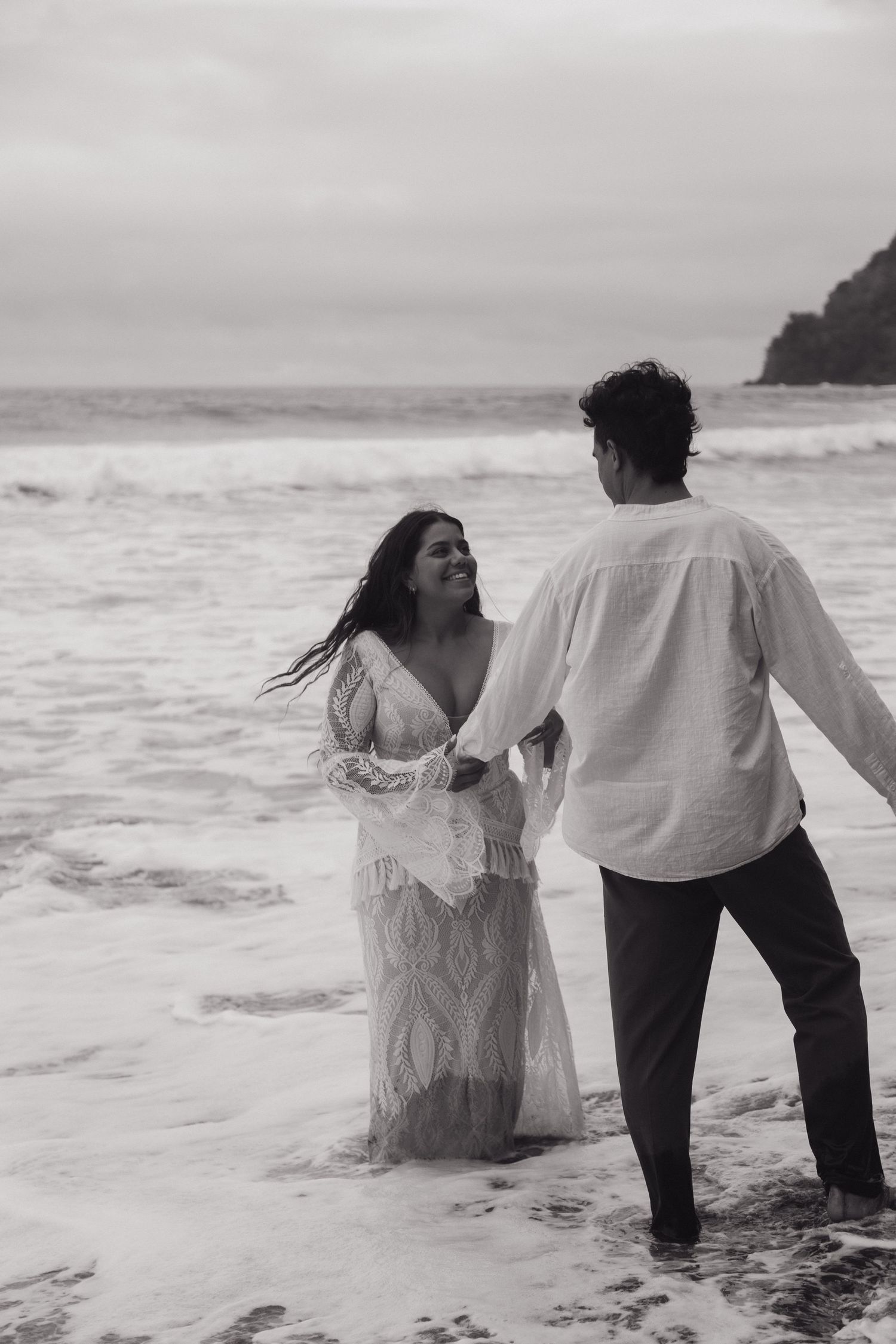 Black and white photo of a couple holding hands in ocean waves, woman in lace dress, romantic beach shoot.