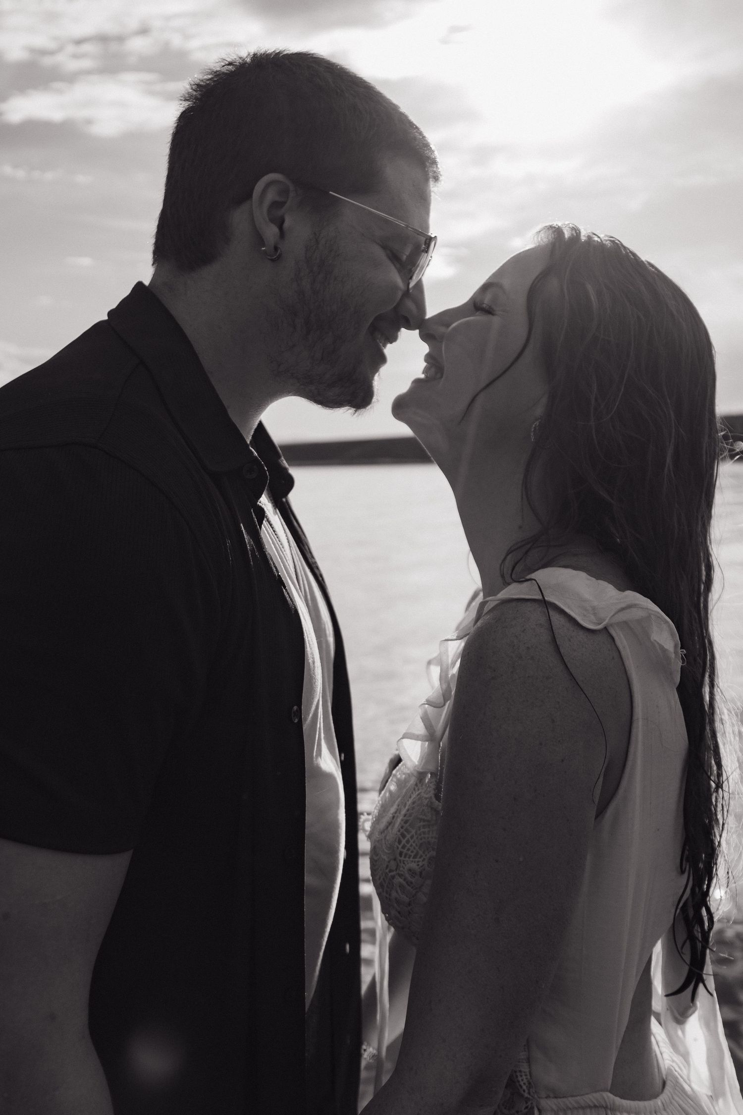 Black and white close-up of smiling couple nearly kissing near a lake with sunlight behind them.