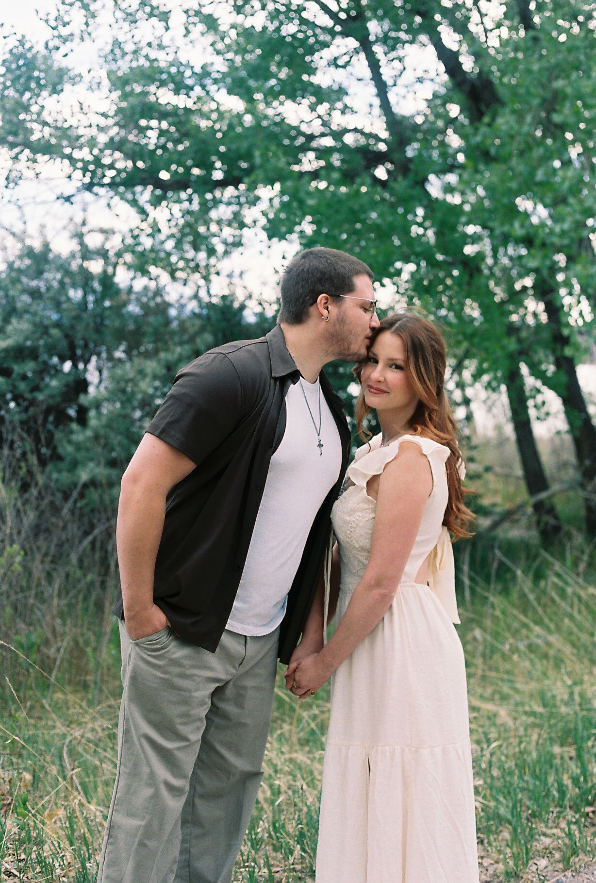 Couple holding hands in wooded area, man kissing woman's forehead in white summer dress.