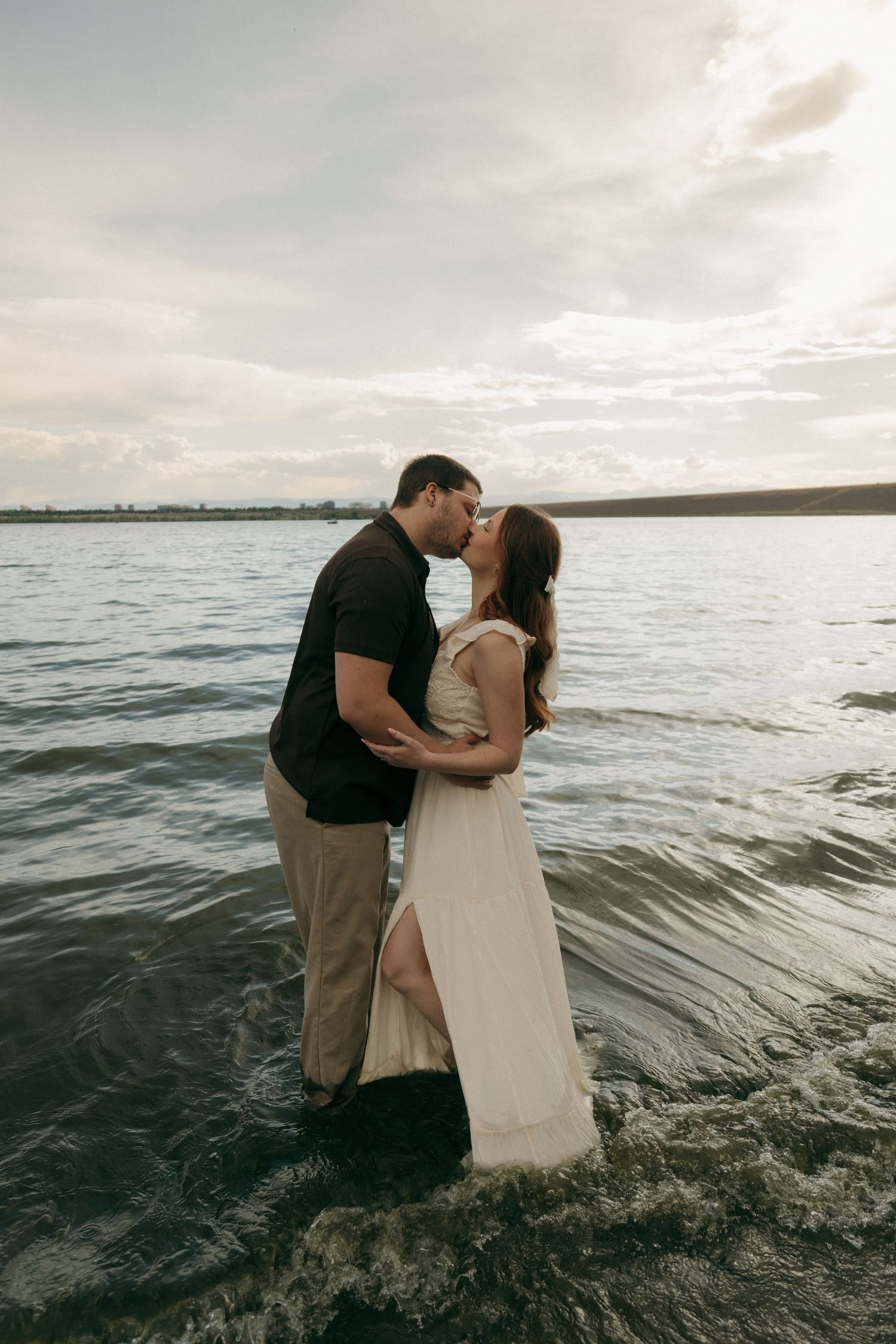 Romantic couple sharing a kiss while standing in shallow lake water at sunset.