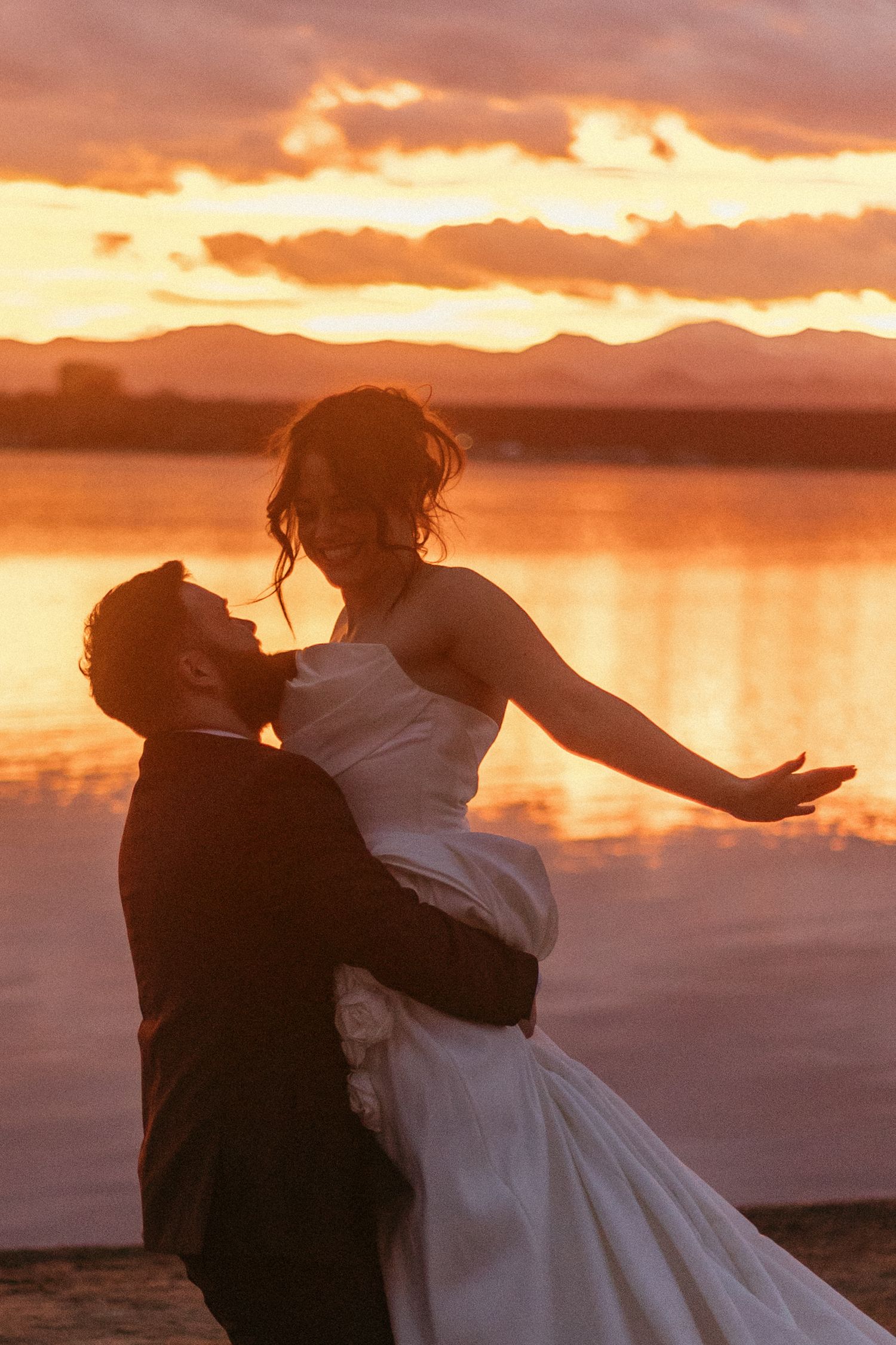 A romantic sunset silhouette of newlyweds embracing by a lake with mountains in the background.