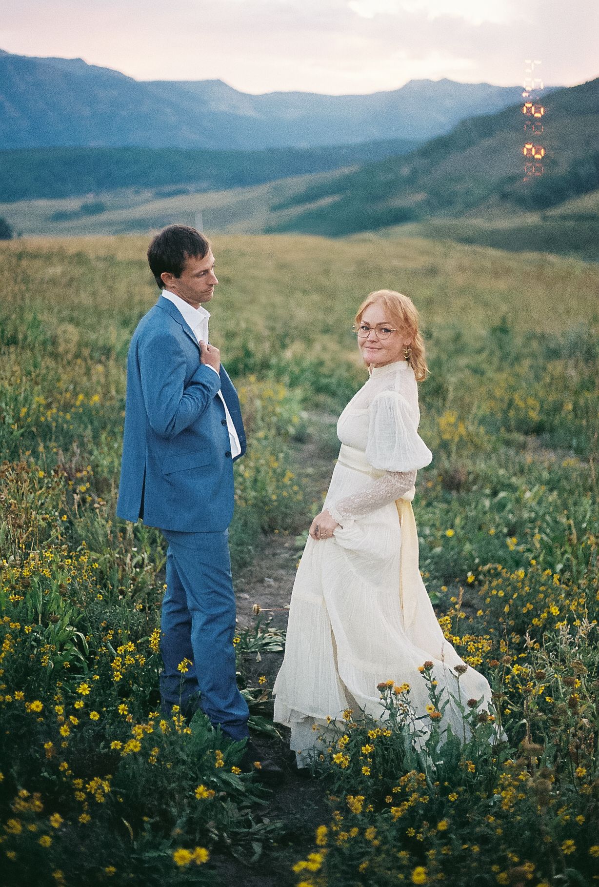 Bride in vintage white dress and groom in blue suit posing in yellow wildflower mountain meadow.