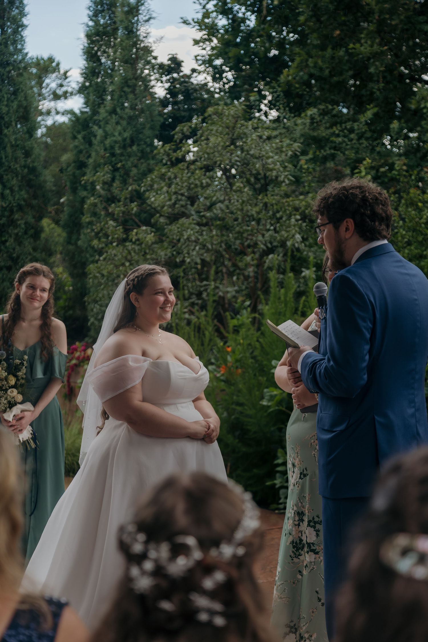 Bride in off-shoulder white gown smiles as groom reads vows at outdoor garden wedding ceremony.